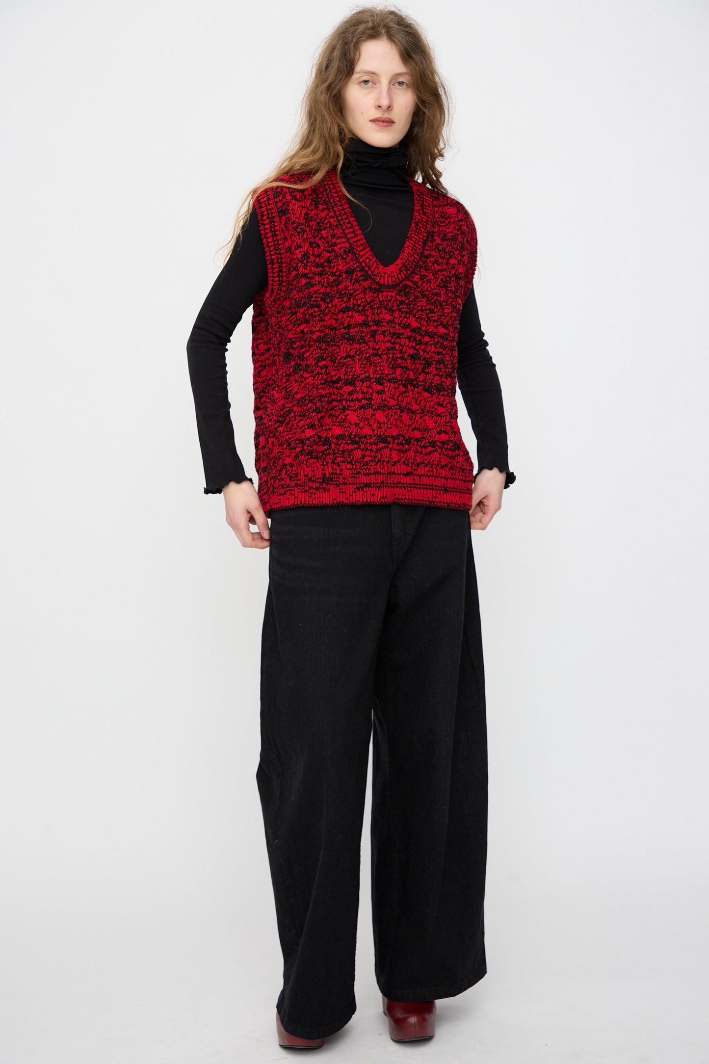 A person with long hair wears a black turtleneck, wide-leg black pants, red shoes, and the Rachel Comey Mati Vest in Red Marble, standing against a plain white background.