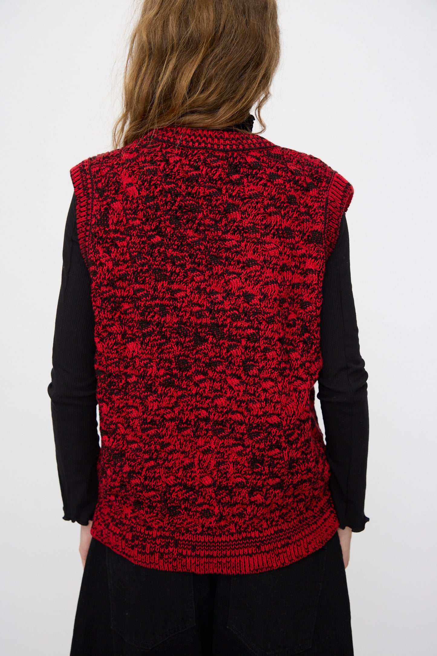 A person with long brown hair faces away, wearing a black long-sleeve shirt and the Rachel Comey Mati Vest in Red Marble, featuring a relaxed fit and bold patterned design.