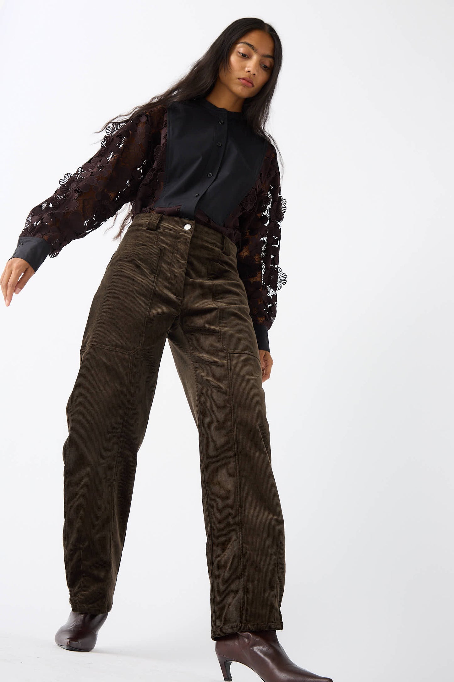 A person with long hair wearing a black blouse with sheer sleeves and high-waisted Rachel Comey Mid Wale Corduroy Oberlin Pants in Bark stands against a white background.