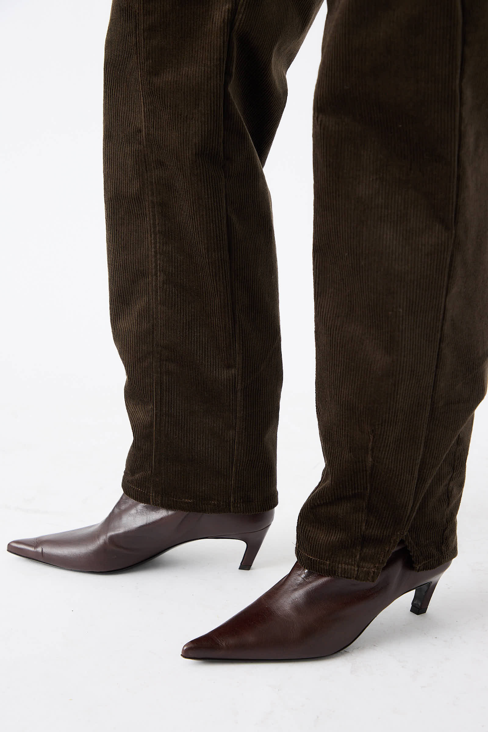 A person wearing dark brown pointed-toe shoes and the high-waist, relaxed-fit Mid Wale Corduroy Oberlin Pant in Bark by Rachel Comey stands against a plain white background.