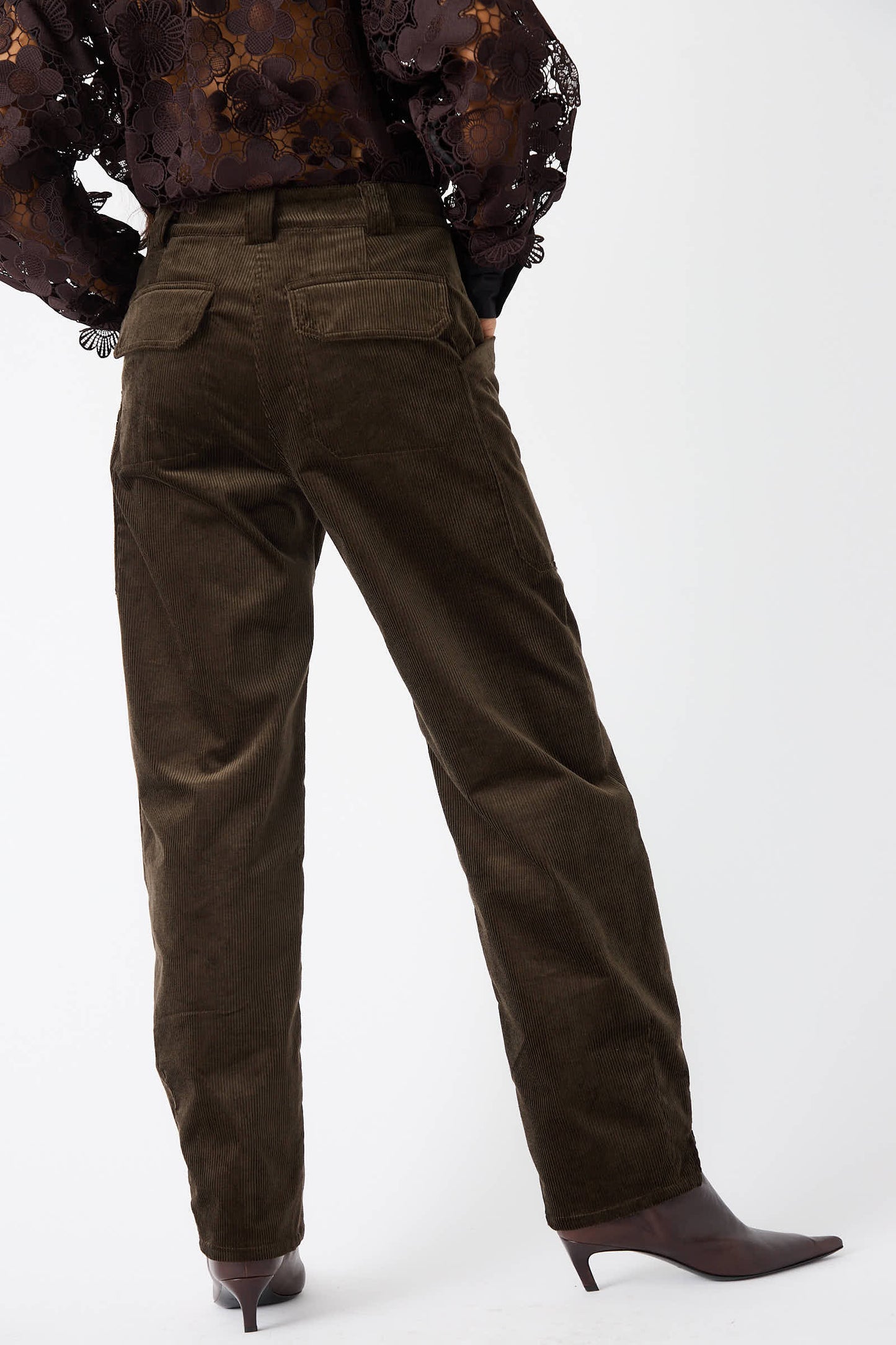 Individual dressed in Rachel Comey’s Mid Wale Corduroy Oberlin Pant in Bark, complemented by a long-sleeve lace top and brown heeled shoes, standing with their back to the camera.