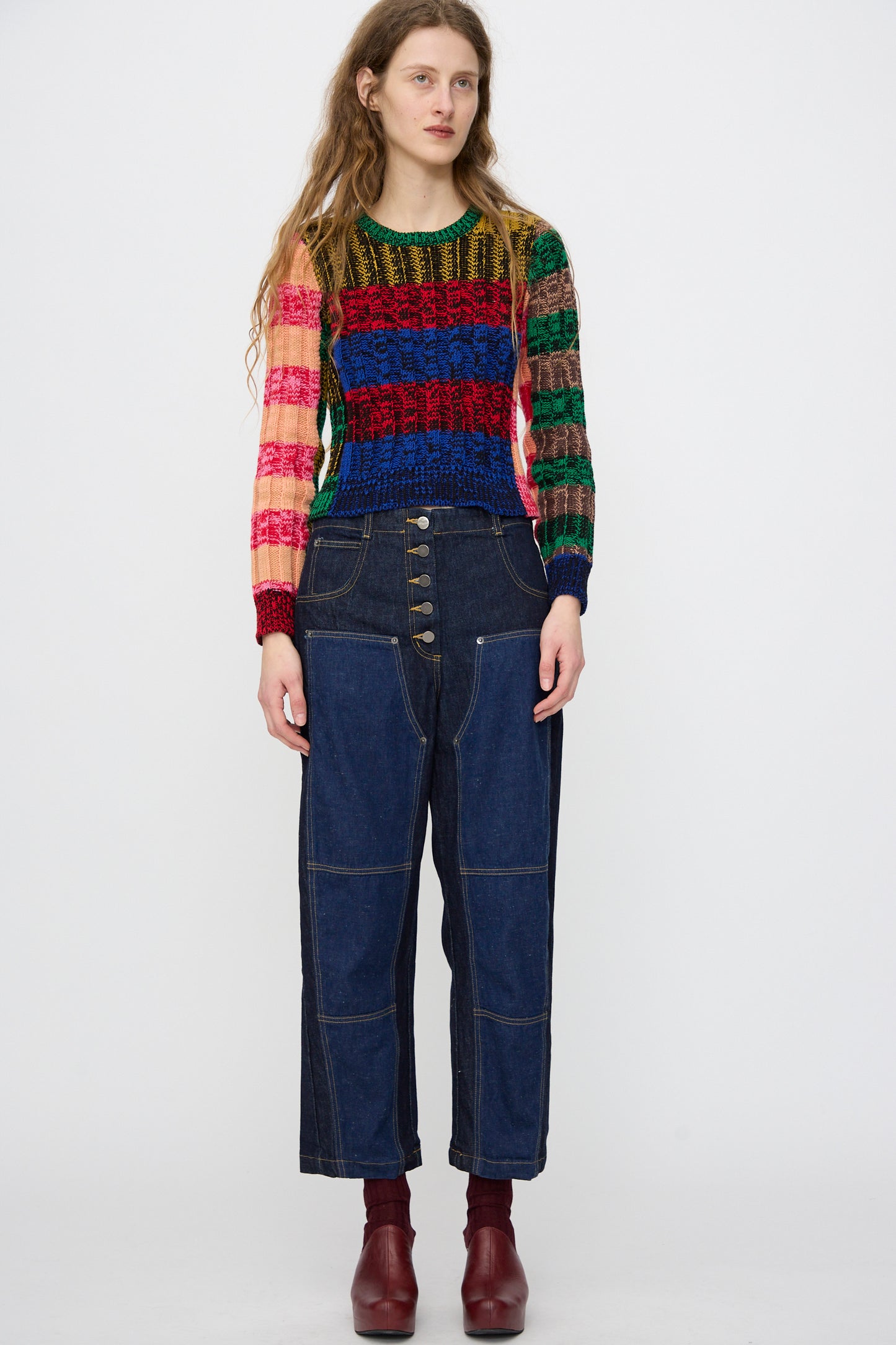 A person stands against a white background wearing the Rachel Comey Muna Pullover in Stripe Multi, paired with high-waisted dark denim jeans with prominent buttons and maroon ankle boots.