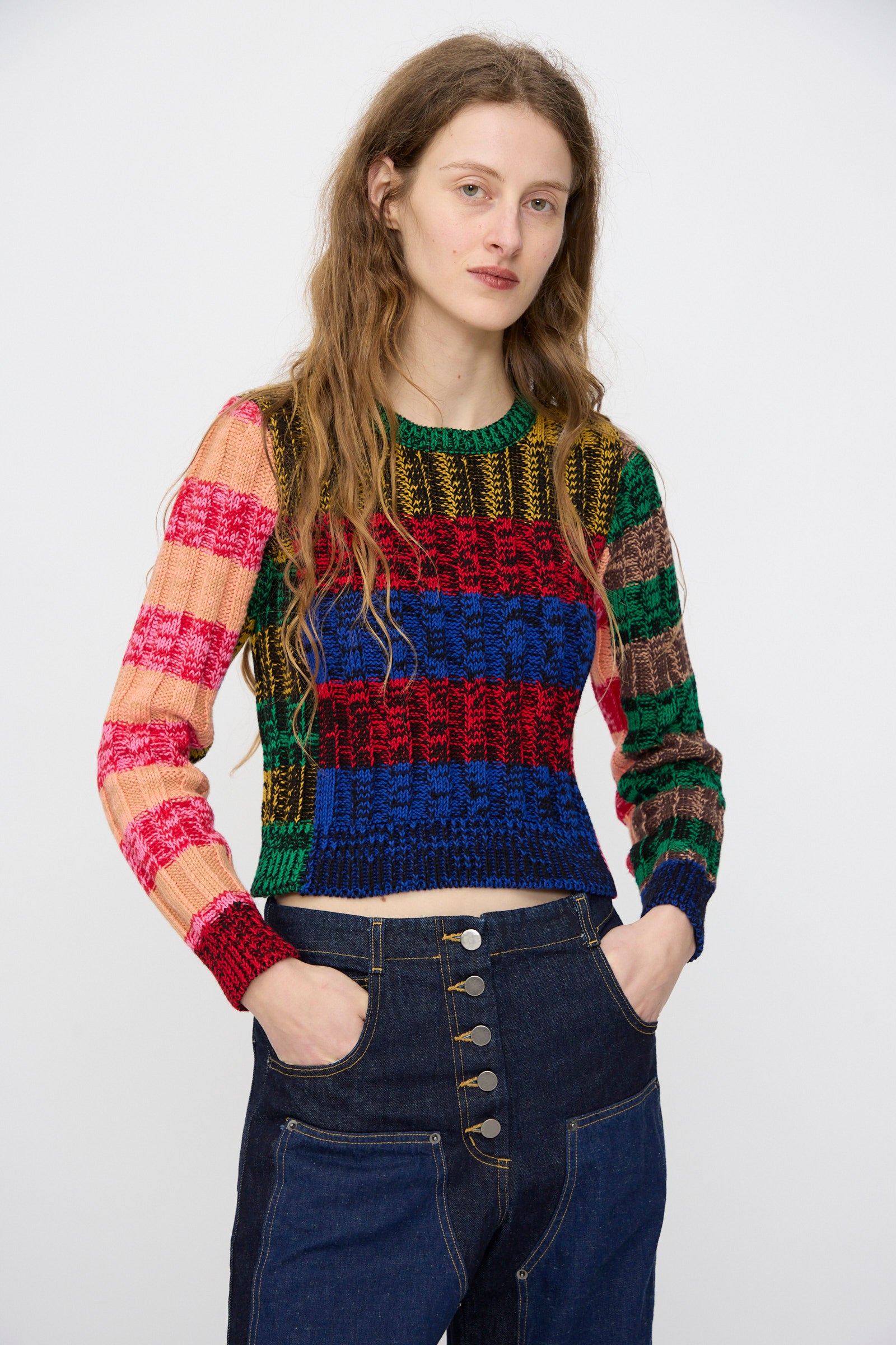 Wearing the Rachel Comey Muna Pullover in Stripe Multi, a person with long wavy hair pairs it with high-waisted dark blue jeans featuring large buttons, standing against a plain light background.