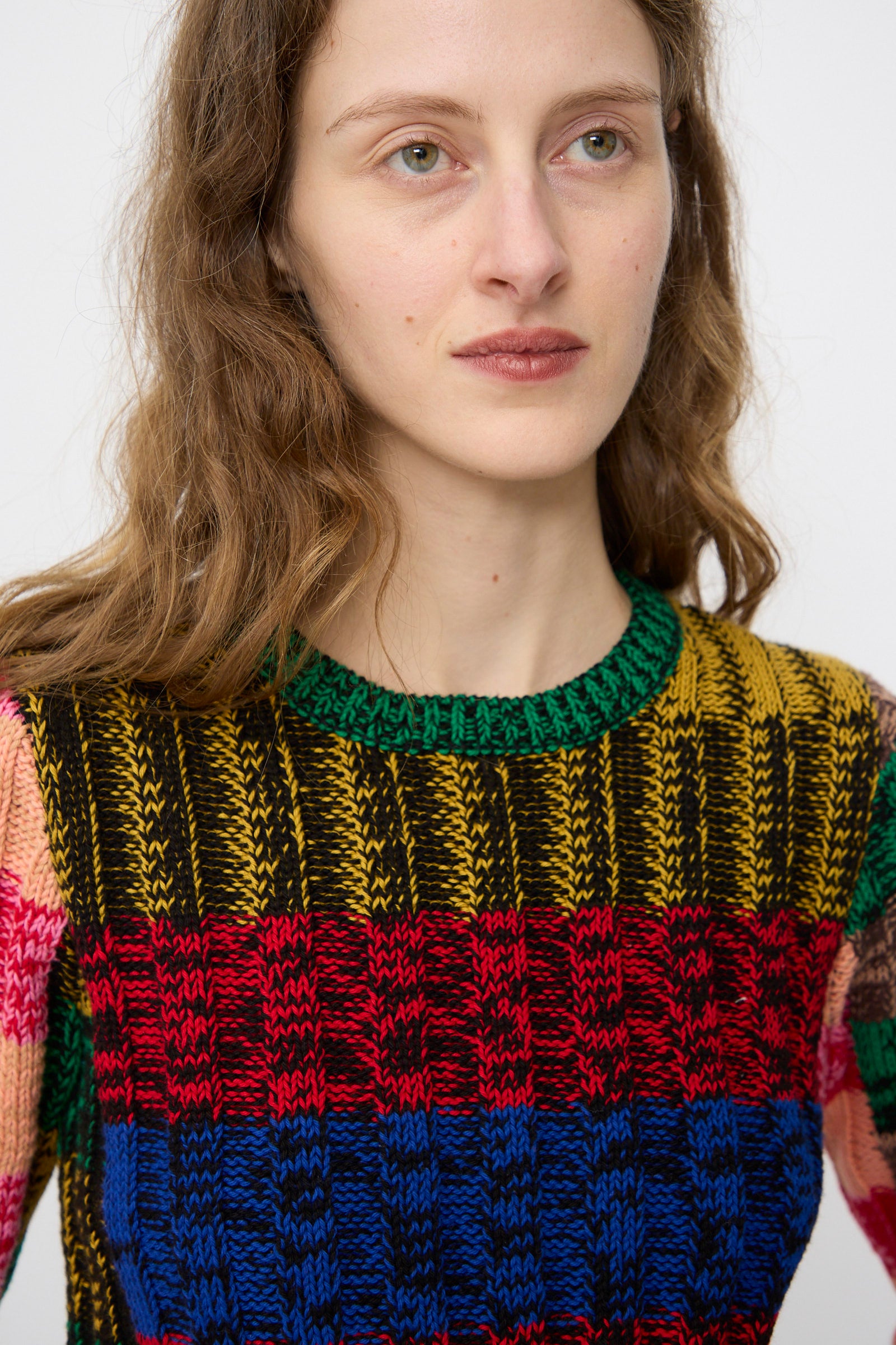 A person with wavy brown hair wears the Rachel Comey Muna Pullover in Stripe Multi—a chunky knit sweater with vibrant yellow, green, red, blue, and black patterns.