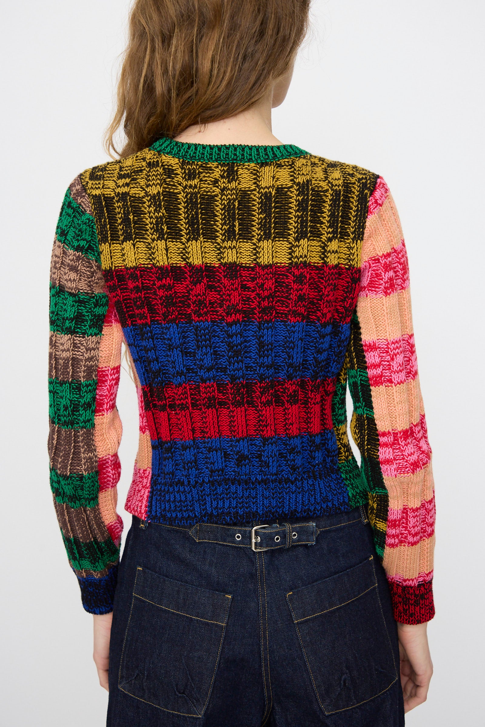 A person is seen from the back wearing the Rachel Comey Muna Pullover in Stripe Multi, a colorful chunky knit sweater with long sleeves and horizontal and vertical stripes, paired with high-waisted dark denim jeans.