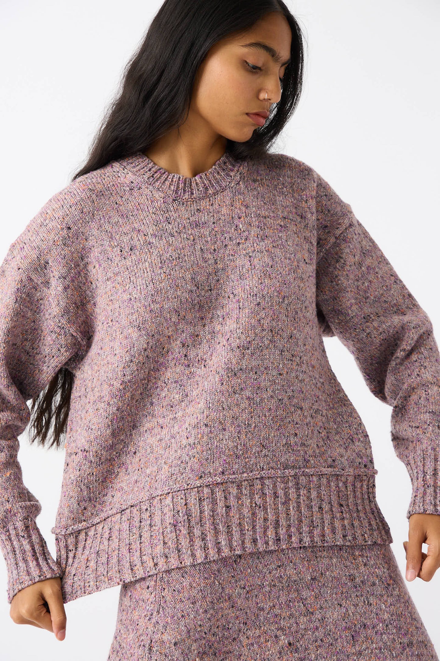 A person with long dark hair wears the Neps Tweed Knit Ninni Sweater in Violet by Rachel Comey, made from recycled cotton, along with a matching skirt. They are looking down to the side against a plain white background.