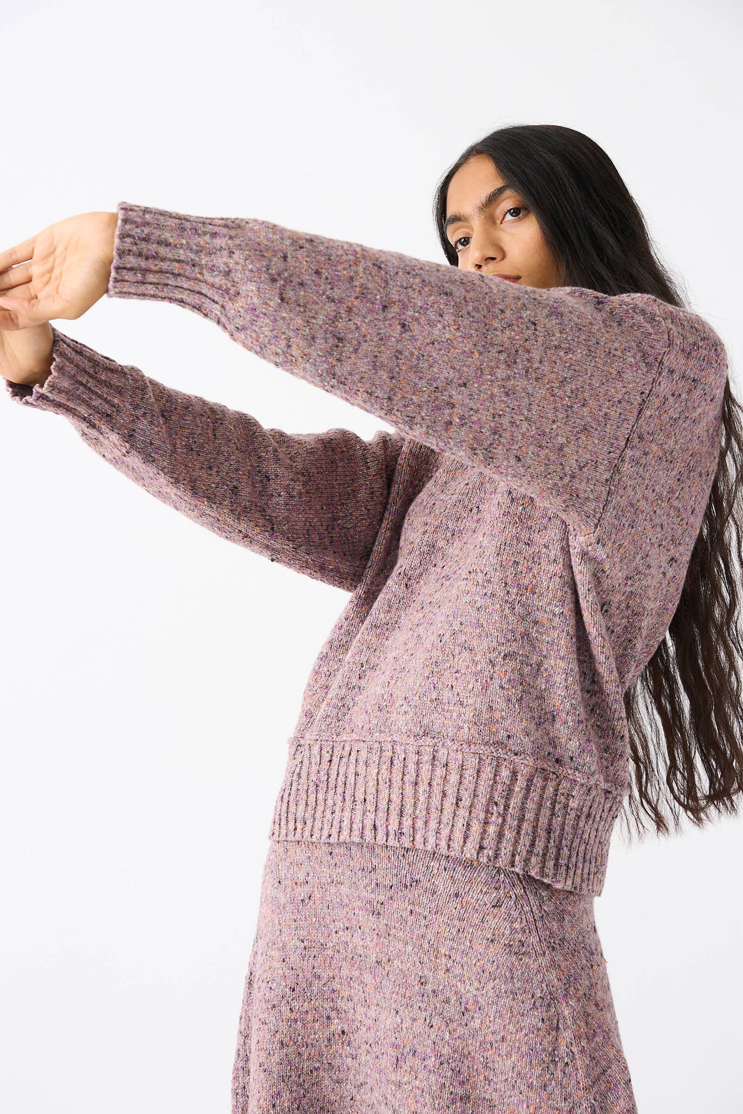 A person with long dark hair, wearing the Rachel Comey Neps Tweed Knit Ninni Sweater in Violet and a matching recycled cotton skirt, extends their arms out in front of them against a plain white background.