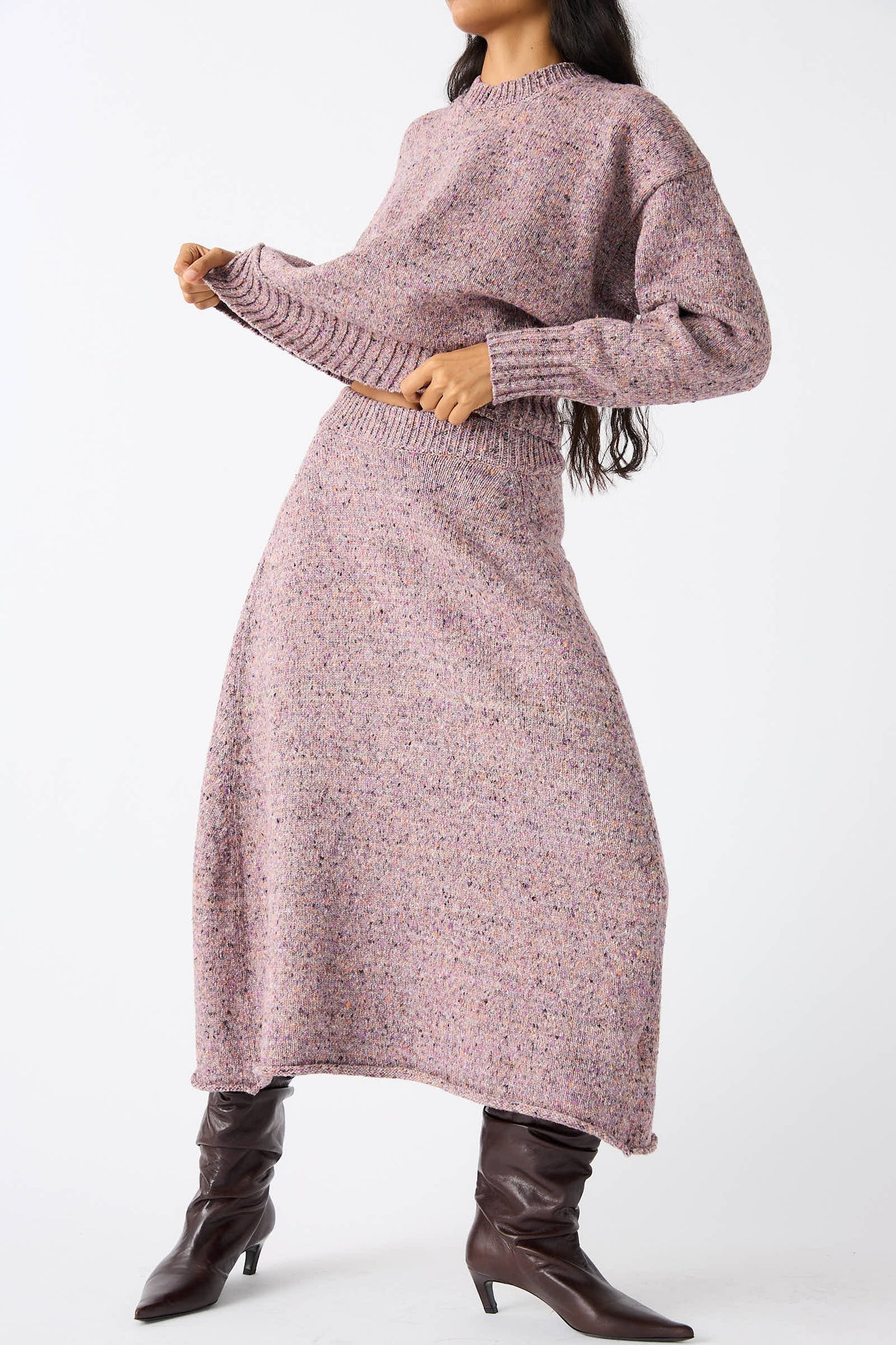 An individual dressed in a Rachel Comey Neps Tweed Knit Rapt Skirt in Violet and a matching long-sleeve sweater, complemented by dark brown knee-high boots, is slightly lifting the hem of their sweater.