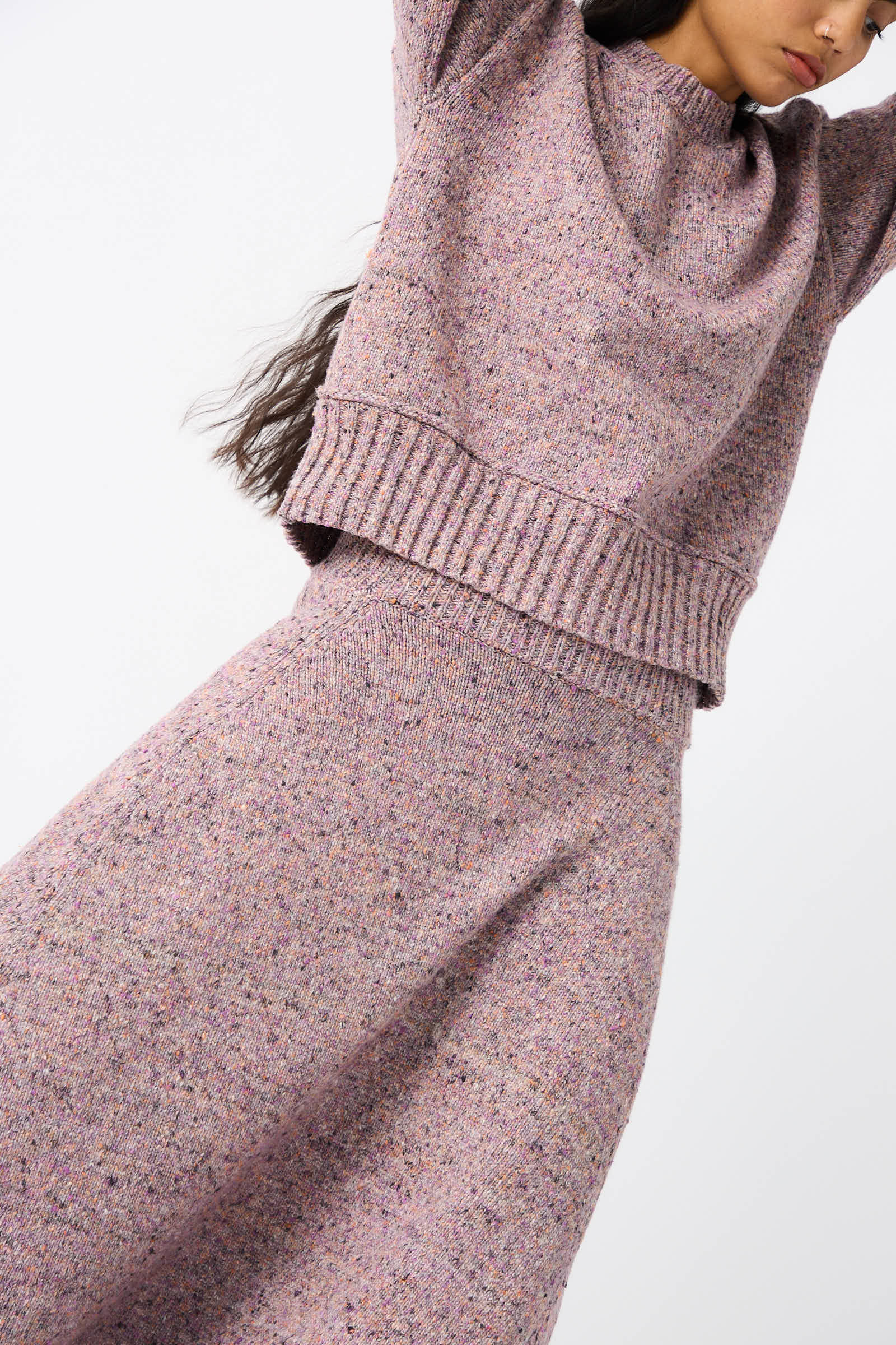 A person dressed in the Rachel Comey Neps Tweed Knit Rapt Skirt in Violet. The light purple, high-waisted midi skirt boasts a cocoon-shaped design. Their arms are raised above their head, and the photo is cropped to highlight their upper body and midsection.