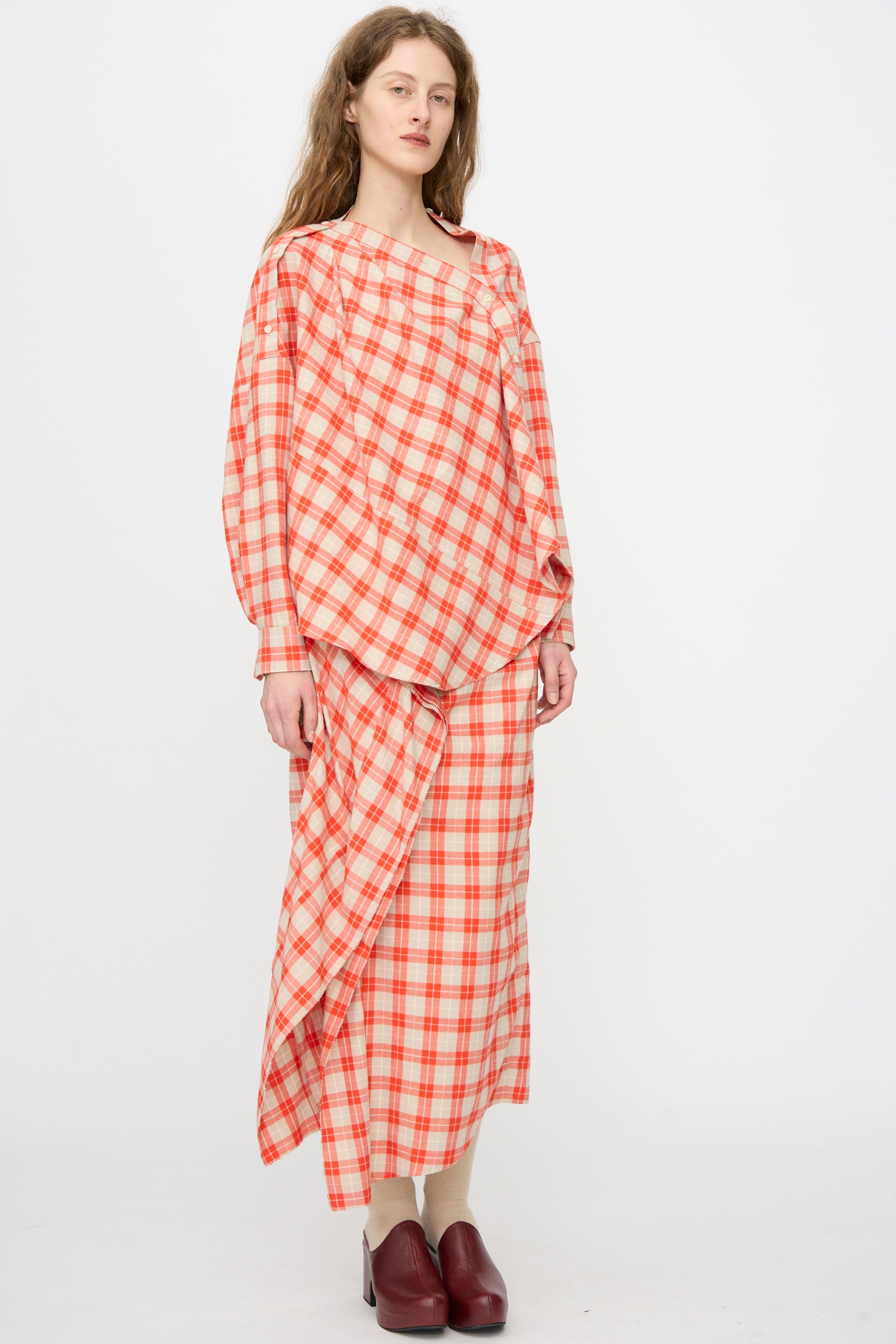 A woman stands against a plain background wearing the Rachel Comey Nesom Skirt in Red, a red and white plaid midi with ruffle detail, paired with a matching loose top and maroon shoes.