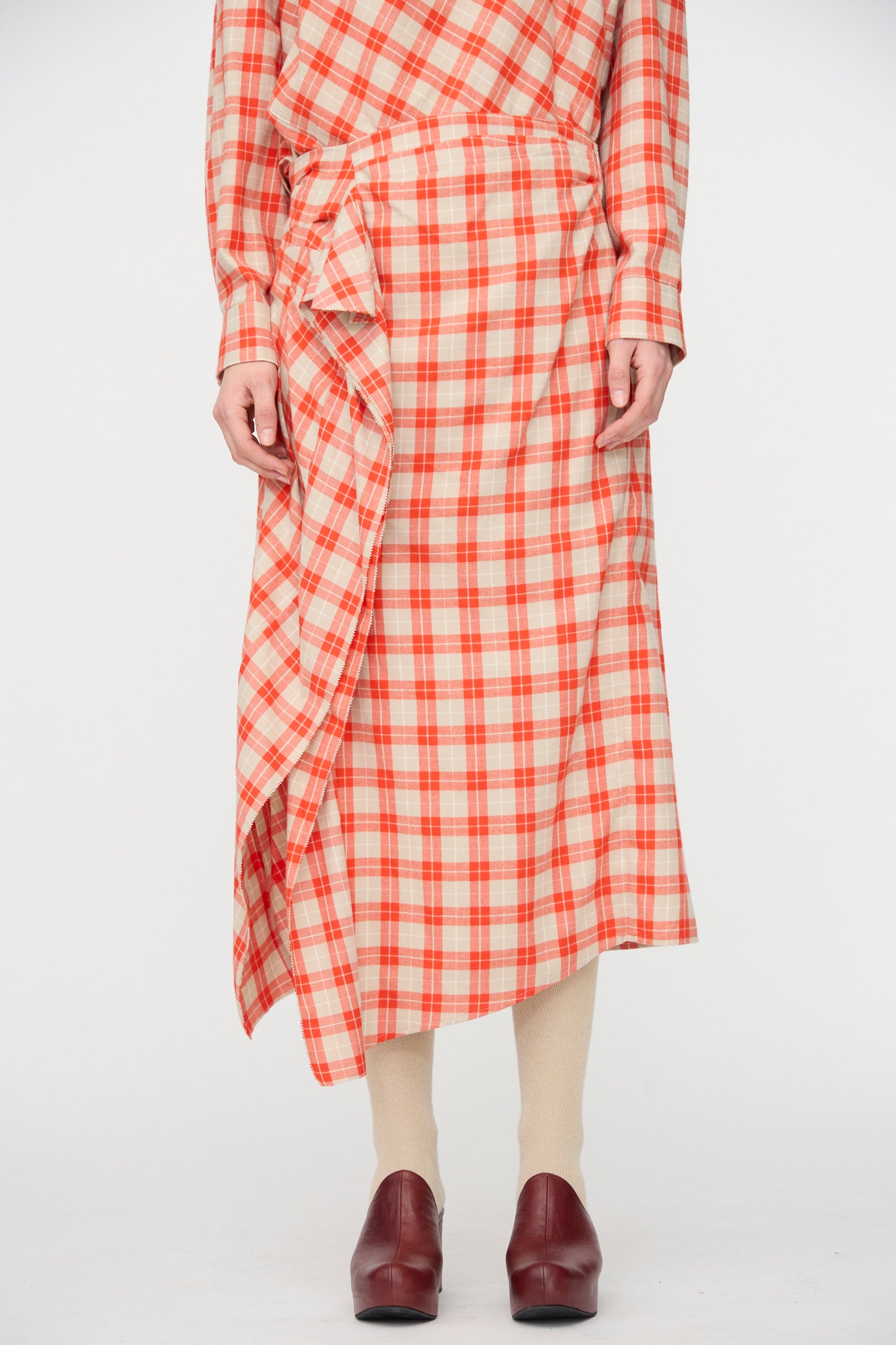 A person wears the Rachel Comey Nesom Skirt in Red, featuring plaid and ruffle details, paired with cream tights and brown shoes, hands resting by their sides.