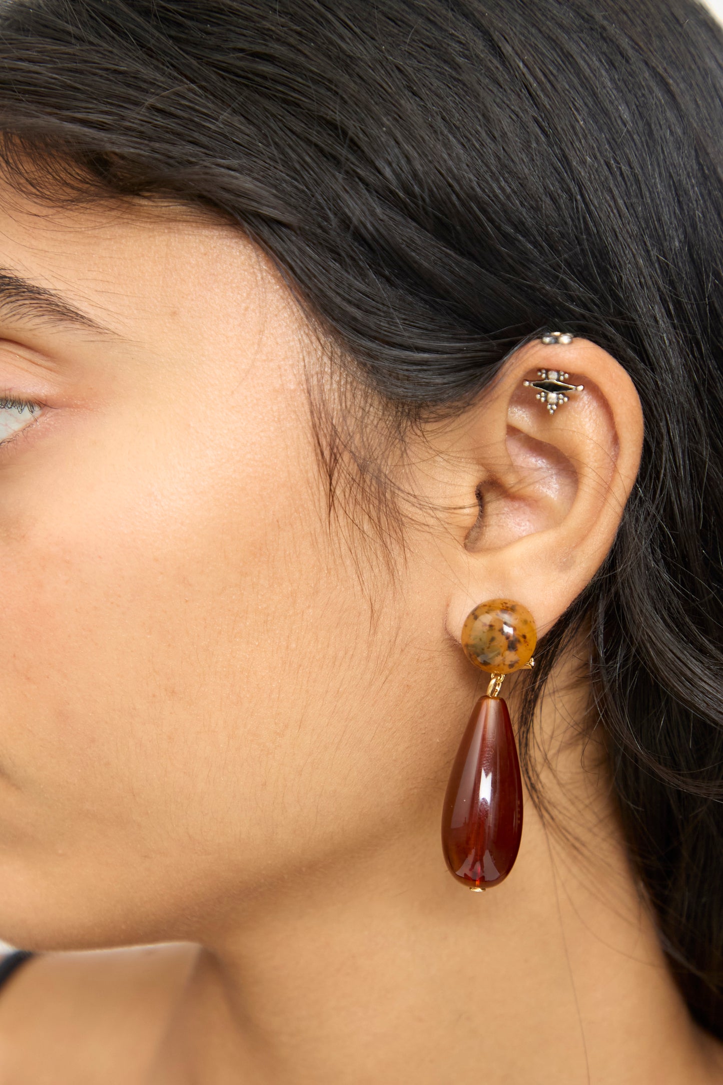 A close-up of a person's ear wearing the Rachel Comey Pamploma Earrings in Picture Jasper Multi—featuring a large, sculptural design with a brown round Picture Jasper stud and a teardrop-shaped pendant—alongside a small barbell piercing in the upper ear. This distinctive piece is made in Italy.