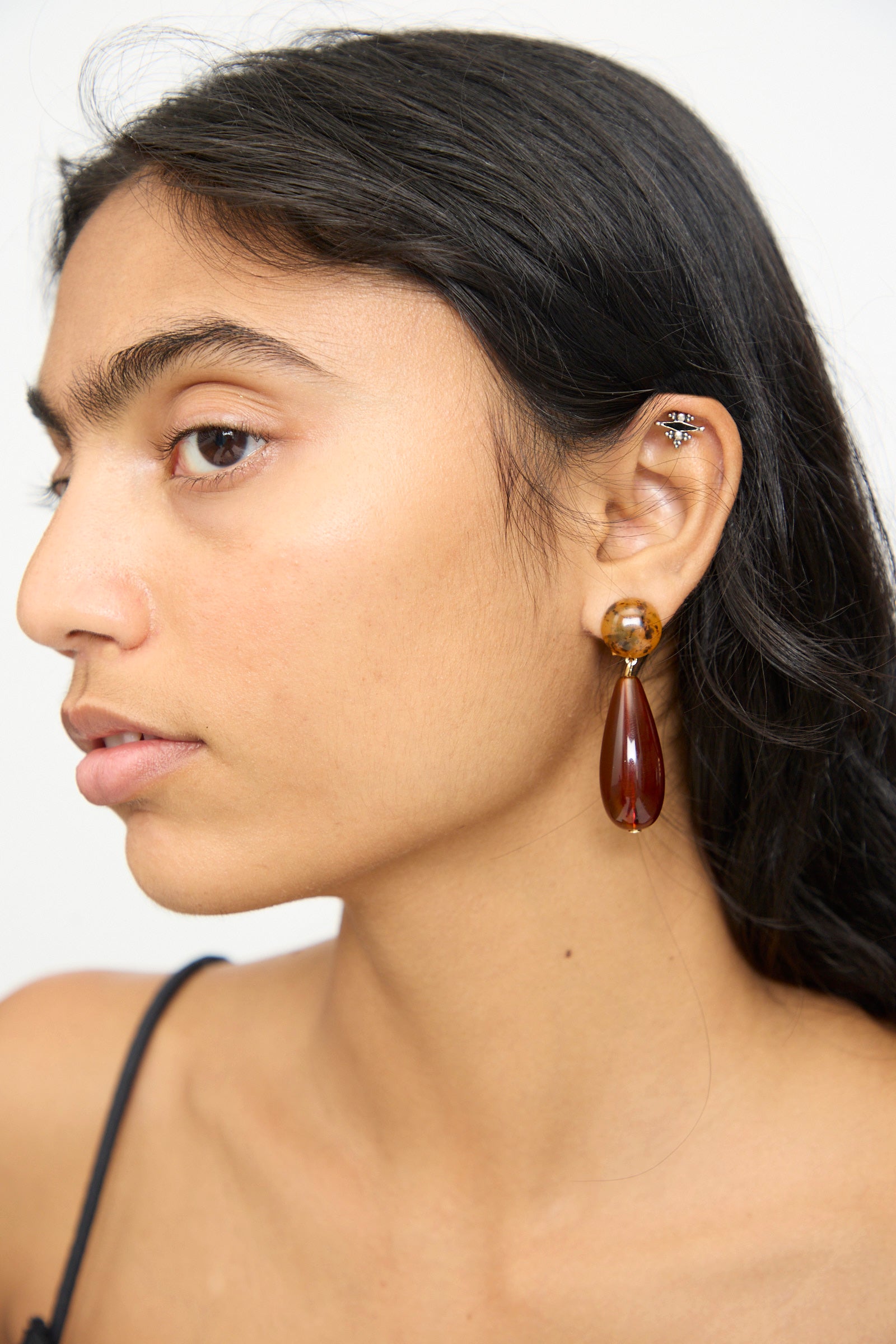 Woman with long dark hair wearing the Rachel Comey Pamploma Earrings in Picture Jasper Multi—a sculptural, dark teardrop-shaped earring—along with multiple small silver ear piercings, looking to the left against a light background.