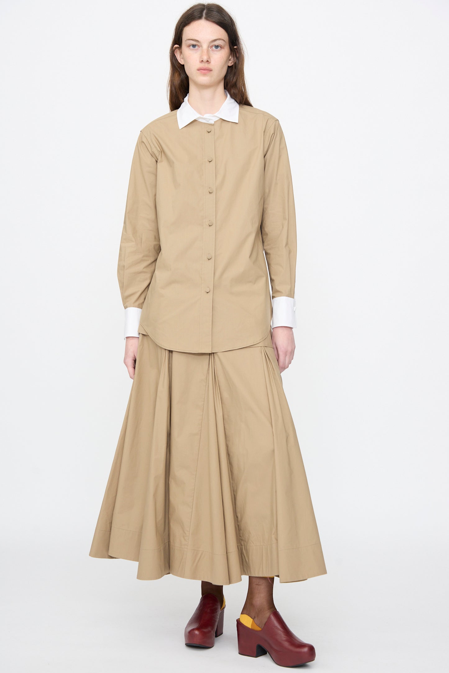 A woman stands against a plain background wearing the Rachel Comey Pierre Shirt in Khaki, styled with a matching pleated skirt and dark red platform shoes featuring yellow heels.
