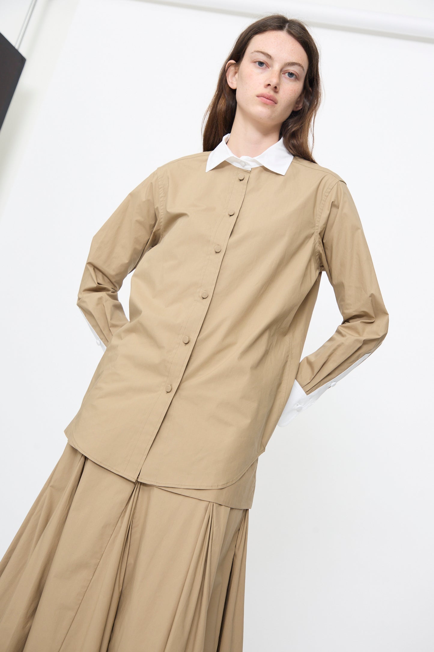 A person wears the Rachel Comey Pierre Shirt in Khaki, a tailored button-up with a white collar, paired with a matching skirt, both crafted from organic cotton, standing against a plain white background.