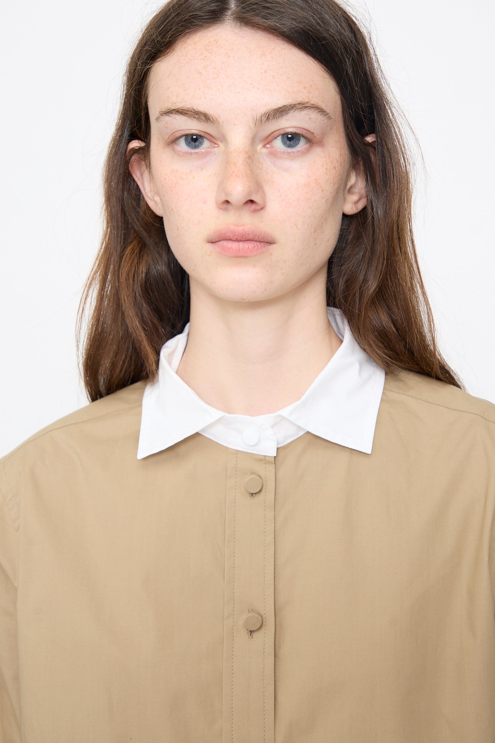 A woman with long brown hair faces forward against a plain light background, wearing the Rachel Comey Pierre Shirt in khaki layered over a white collared shirt.
