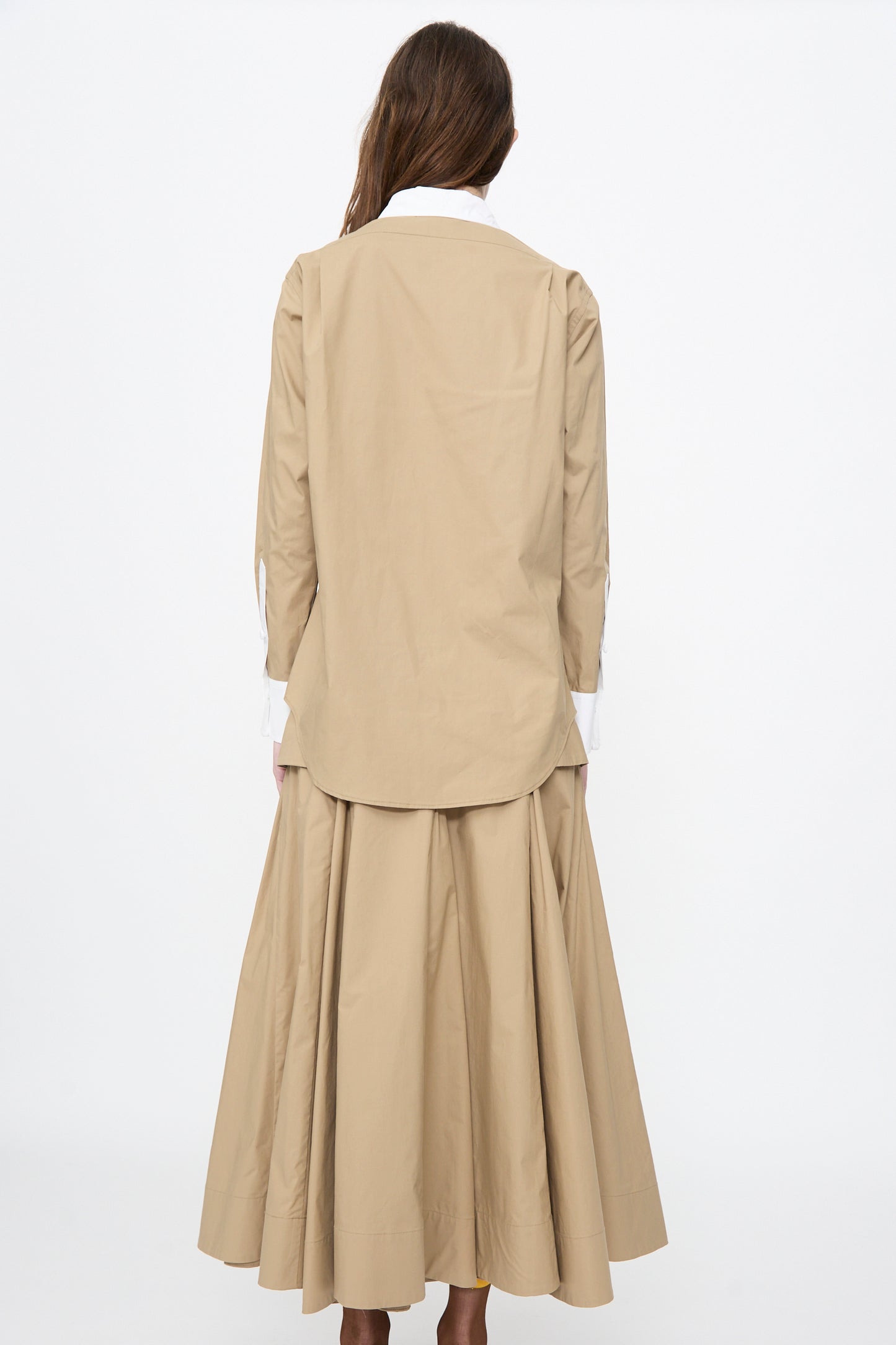A woman stands facing away, wearing the Rachel Comey Pierre Shirt in Khaki with a white collar and cuffs, paired with a matching pleated, ankle-length skirt.