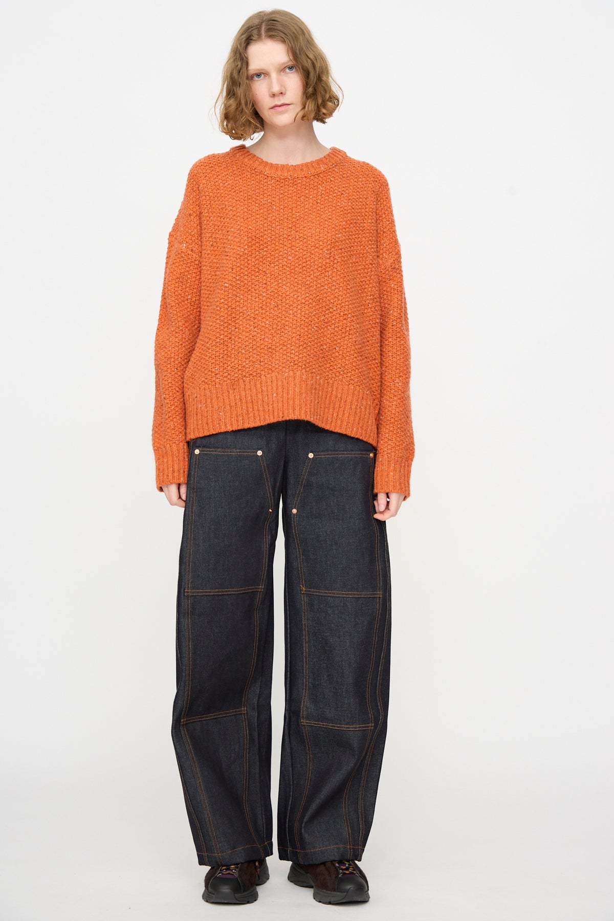 A person stands against a white background wearing the Rachel Comey Prescot Pullover in Orange, paired with loose dark denim jeans featuring visible stitching and dark shoes.