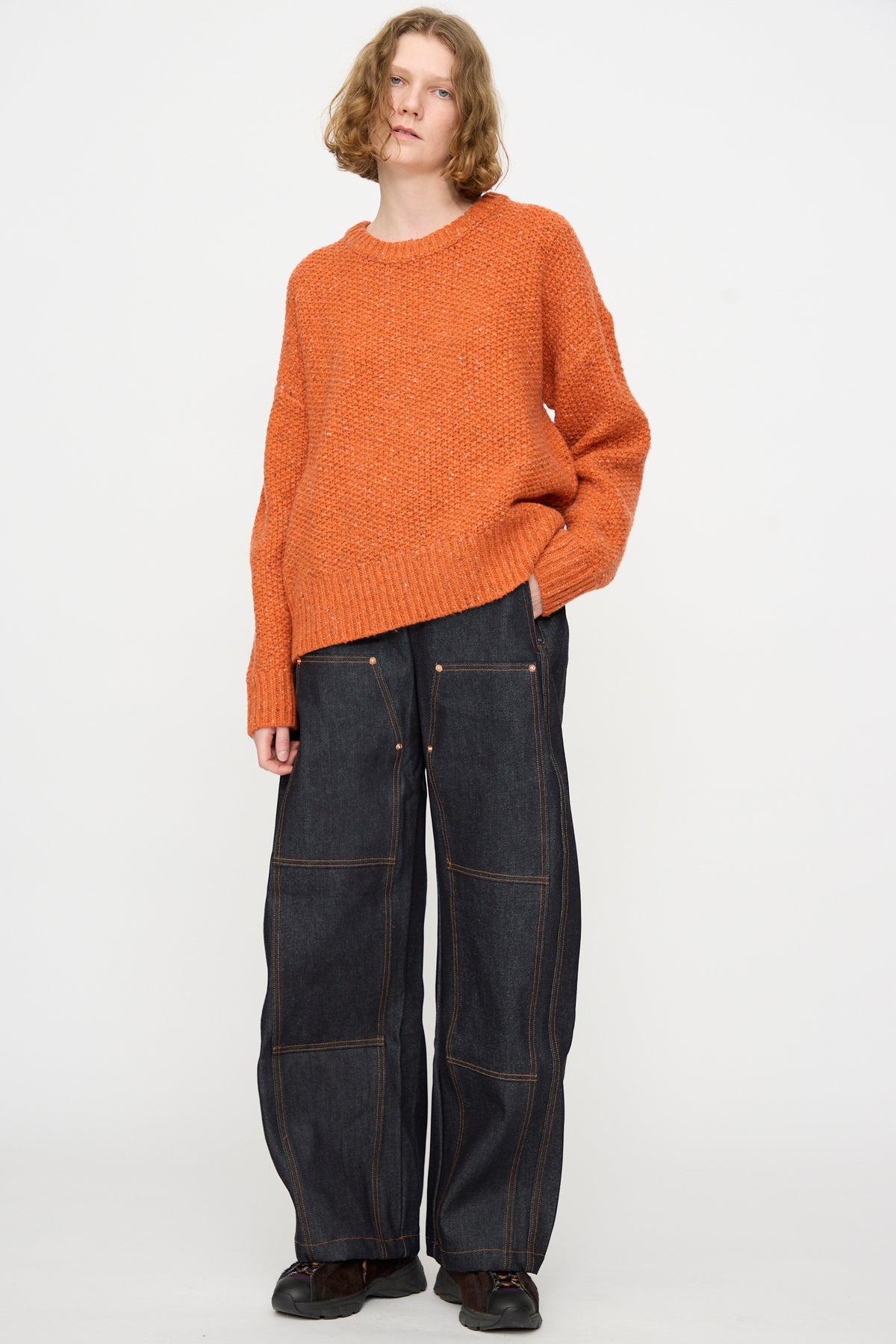 A person wearing the Prescot Pullover in Orange by Rachel Comey and wide-leg dark jeans with visible stitching stands against a plain white background.