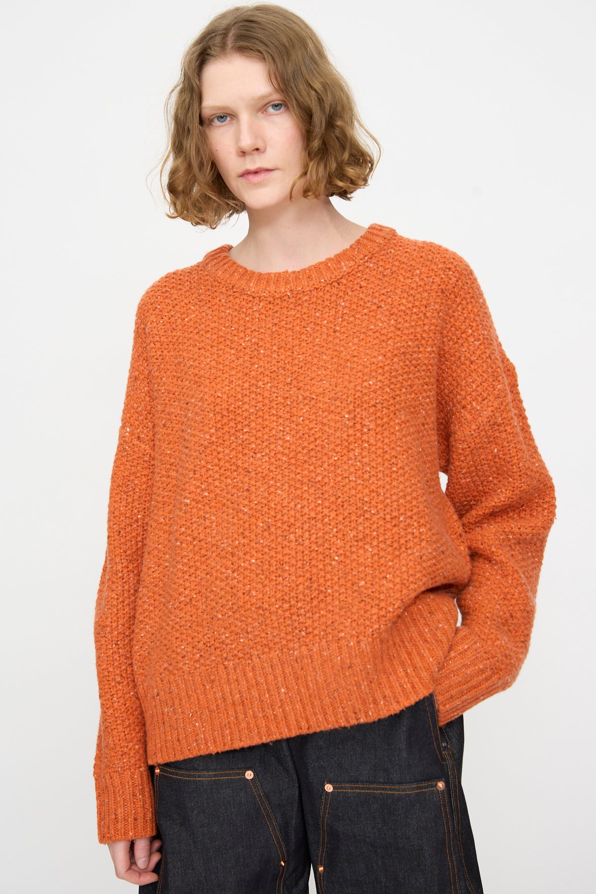 A person with short, wavy hair wears the Rachel Comey Prescot Pullover in Orange and dark jeans, standing against a plain white background.