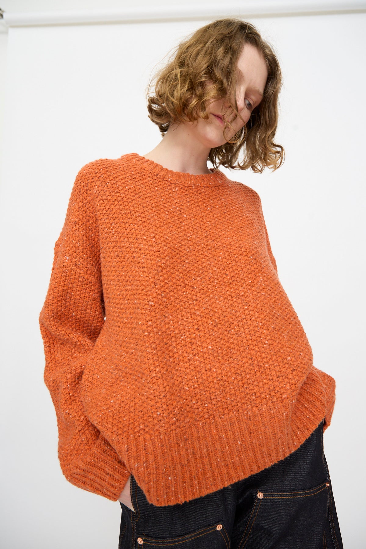 A person wearing the Rachel Comey Prescot Pullover in Orange and dark pants stands against a plain white background, looking downward.