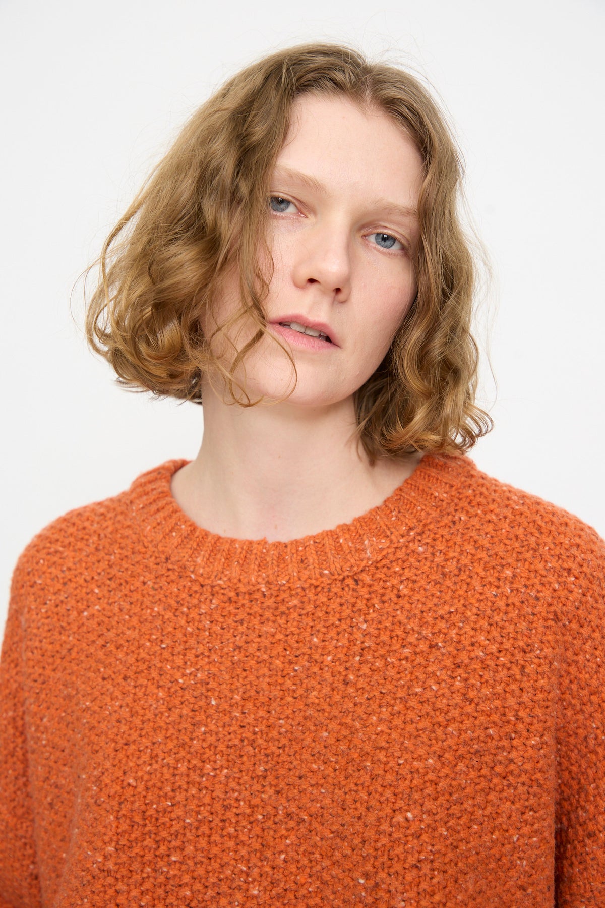 A person with short, wavy light brown hair stands against a plain, light background, wearing the Rachel Comey Prescot Pullover in orange.