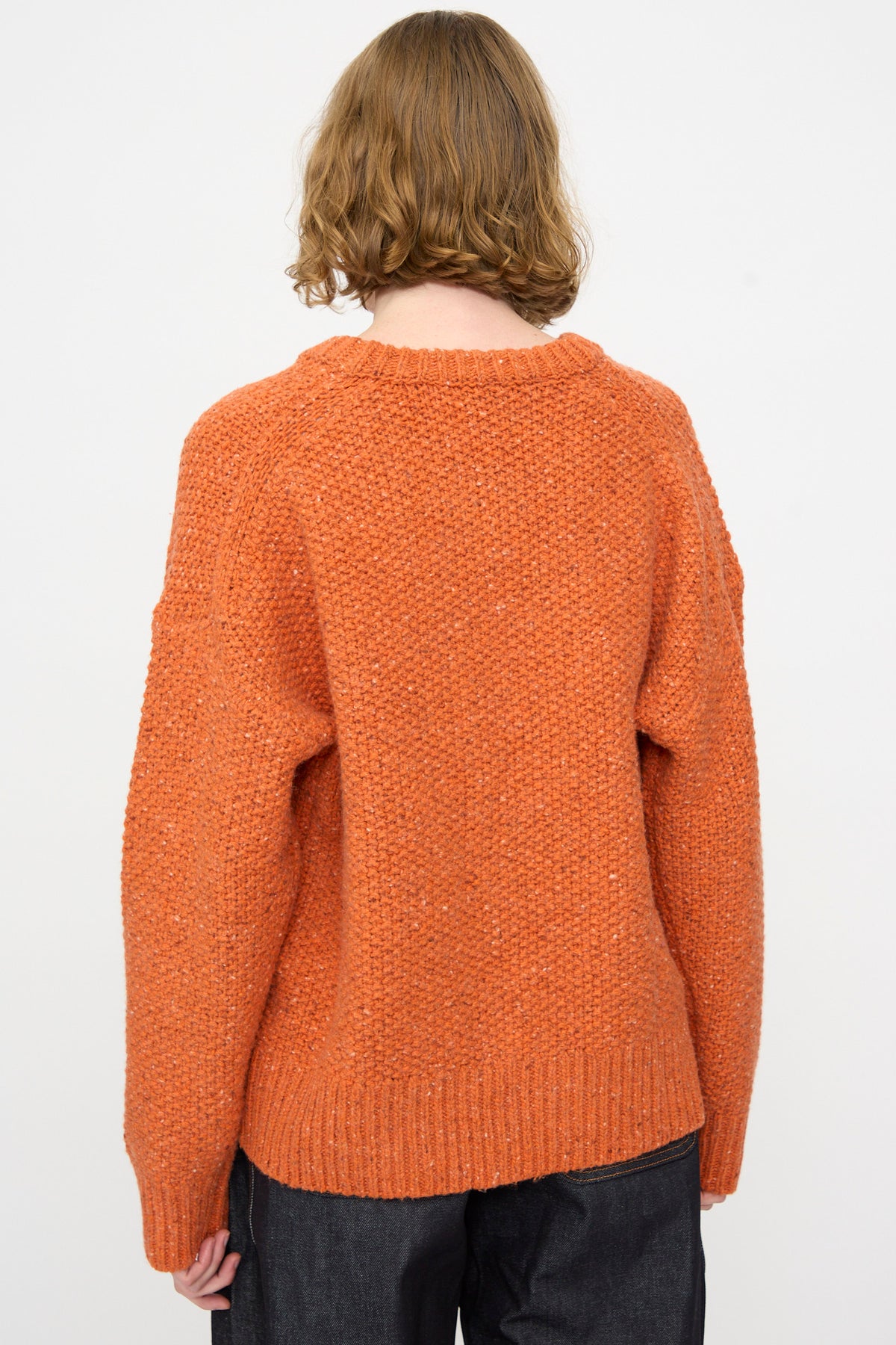 A person with short, wavy light brown hair is shown from the back against a plain white background, wearing the Rachel Comey Prescot Pullover in Orange paired with dark pants.