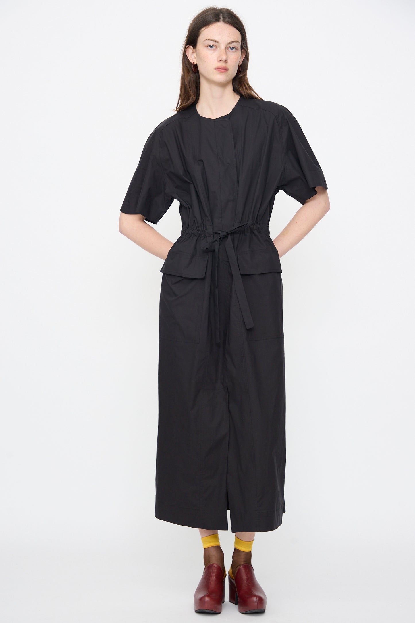 A person wears the Rachel Comey Ramses Dress in Black, a short-sleeve organic cotton midi with large pockets, plus yellow socks and red shoes, standing against a plain white background.