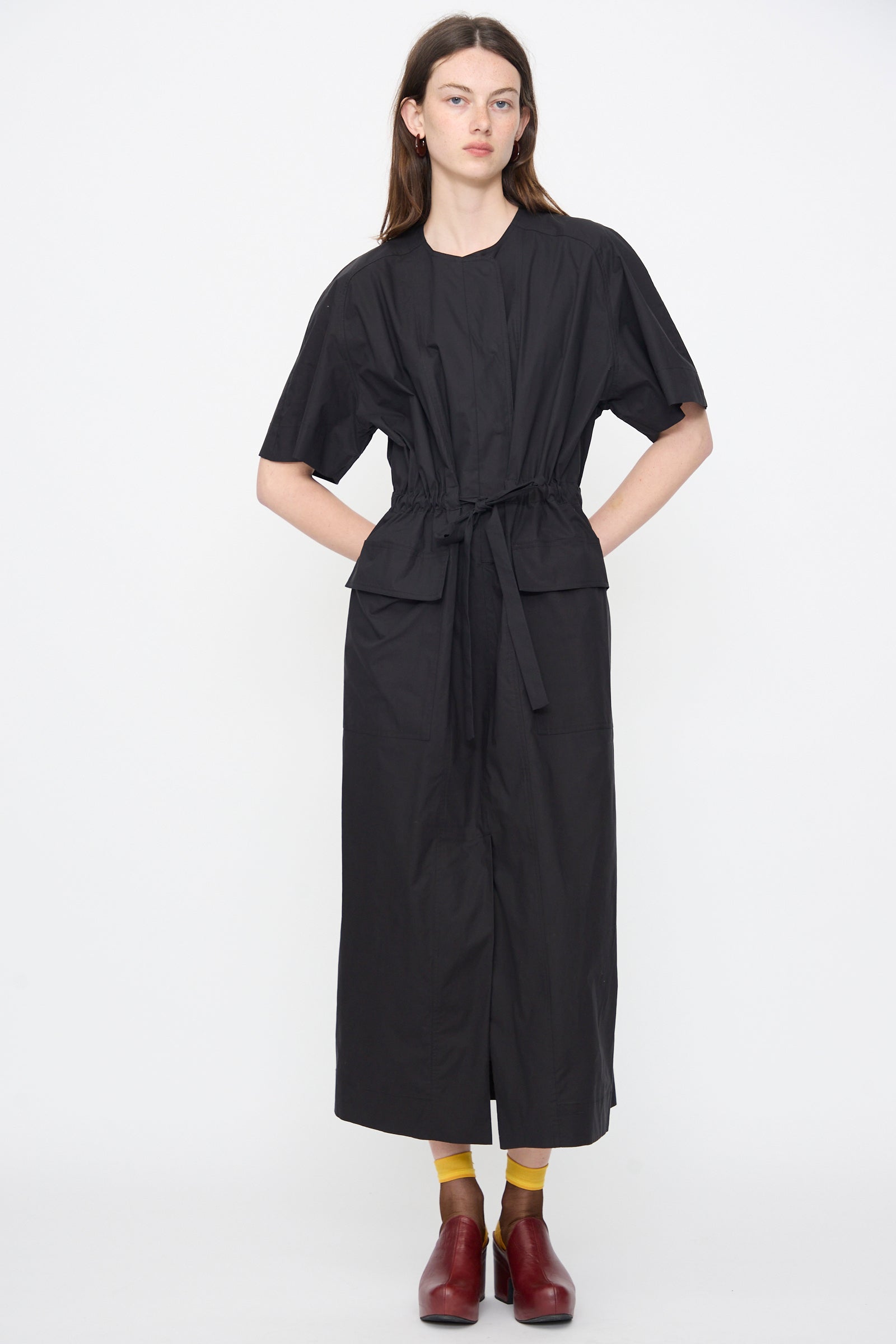 A person wears the Rachel Comey Ramses Dress in Black, a short-sleeve organic cotton midi with large pockets, plus yellow socks and red shoes, standing against a plain white background.