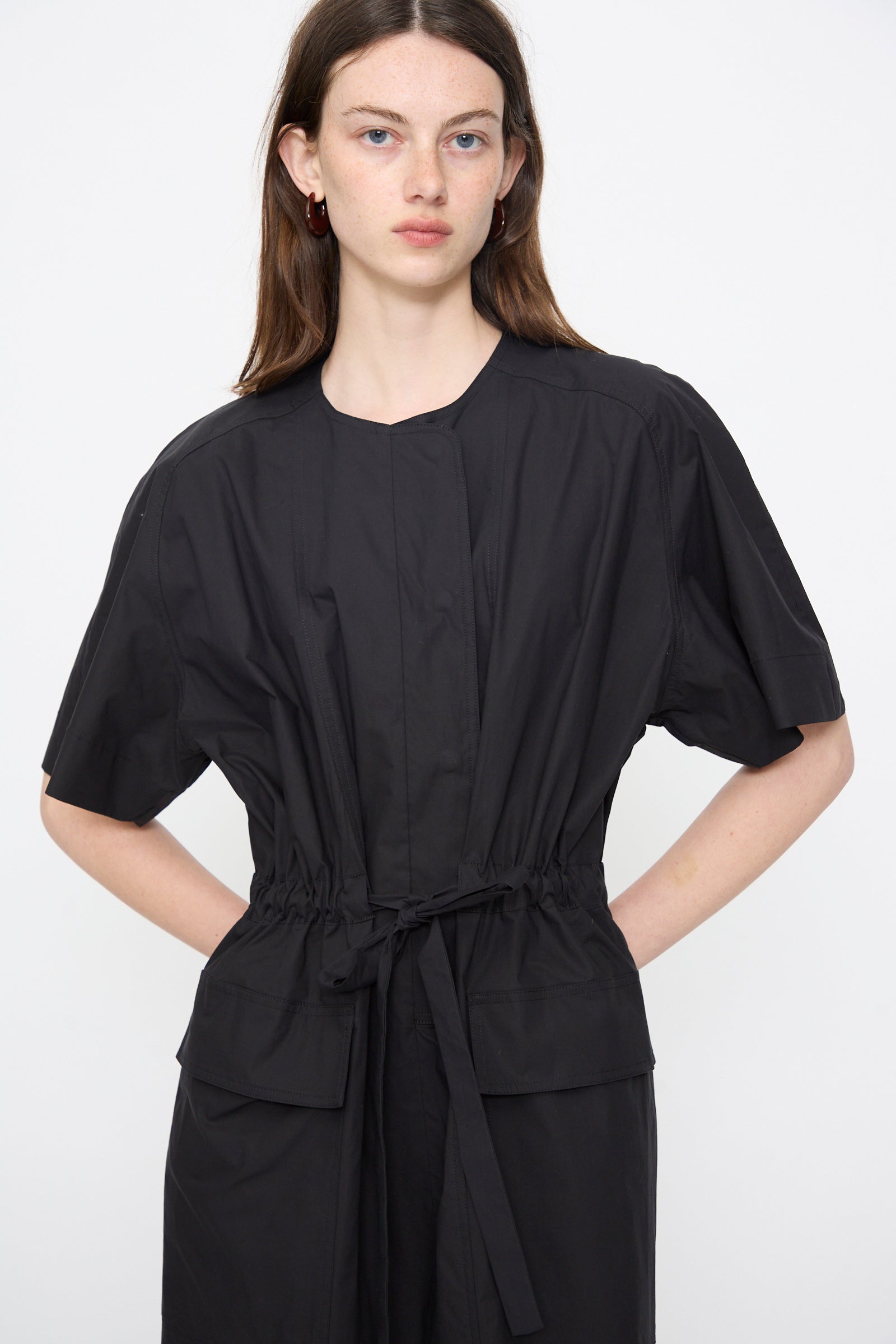 A woman stands against a plain background wearing the Rachel Comey Ramses Dress in Black, a midi dress with short sleeves, two front pockets, and a belted waist, crafted from organic cotton.