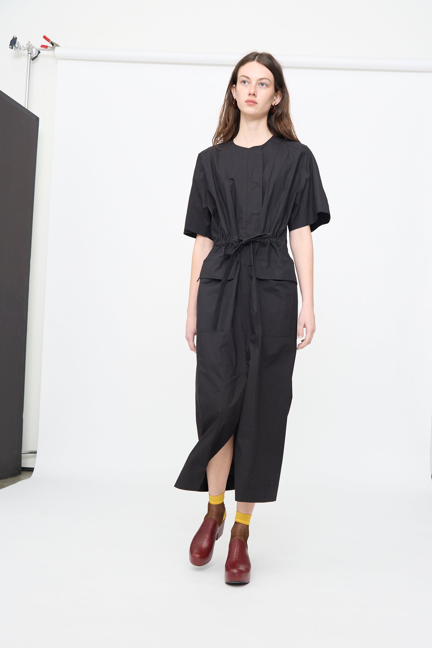 A woman stands in a studio against a plain white background wearing the Rachel Comey Ramses Dress in Black, styled with yellow socks and red shoes.