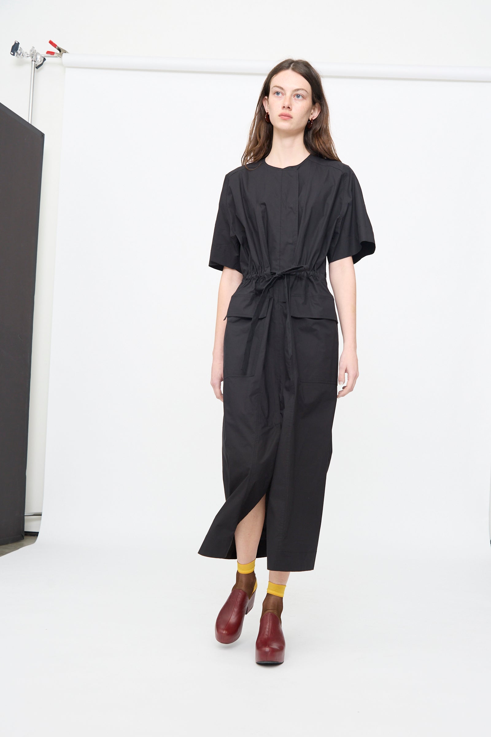 A woman stands in a studio against a plain white background wearing the Rachel Comey Ramses Dress in Black, styled with yellow socks and red shoes.