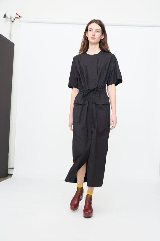A woman stands in a studio against a plain white background wearing the Rachel Comey Ramses Dress in Black, styled with yellow socks and red shoes.