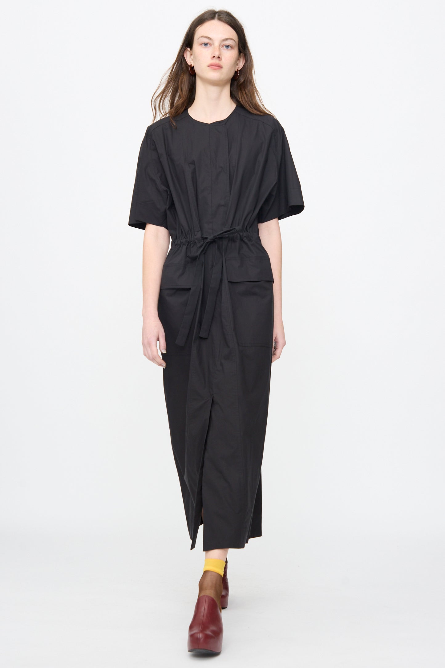 A person stands against a plain background wearing the Rachel Comey Ramses Dress in Black, paired with yellow socks and red shoes.