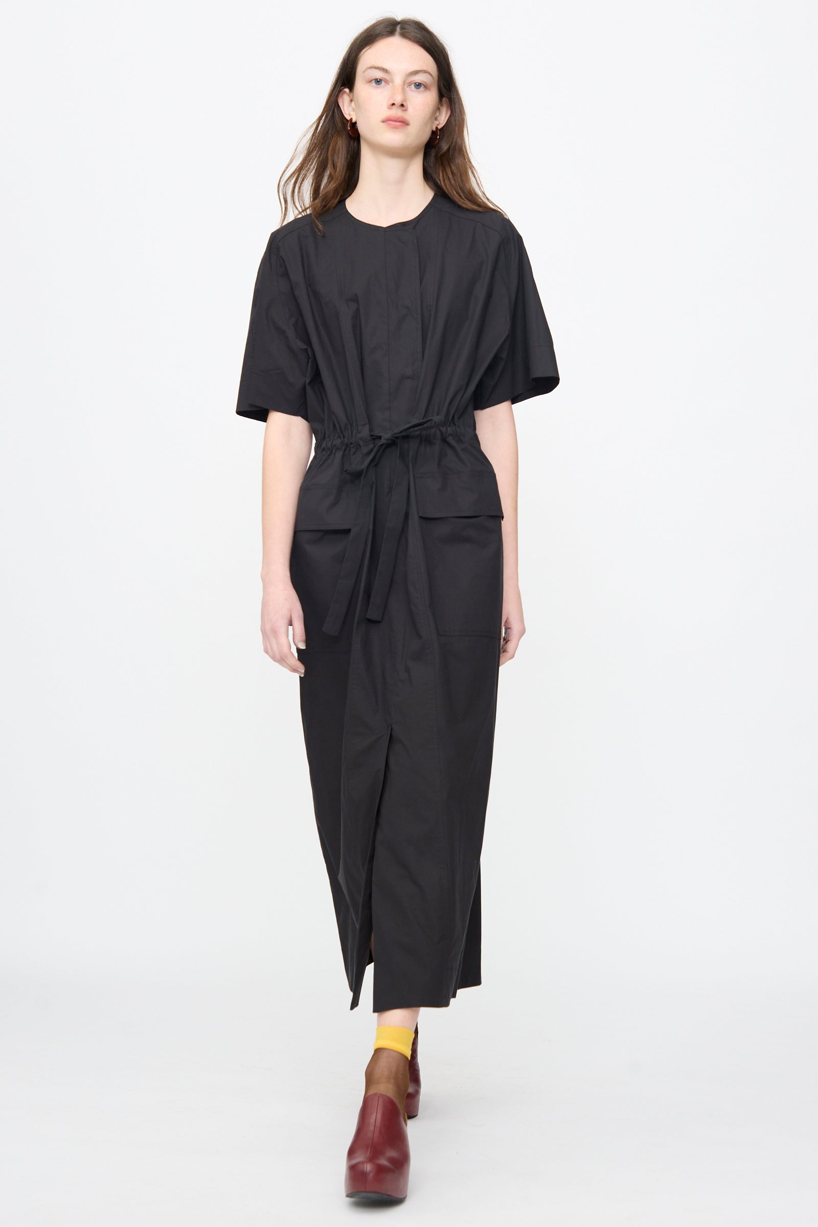A person stands against a plain background wearing the Rachel Comey Ramses Dress in Black, paired with yellow socks and red shoes.
