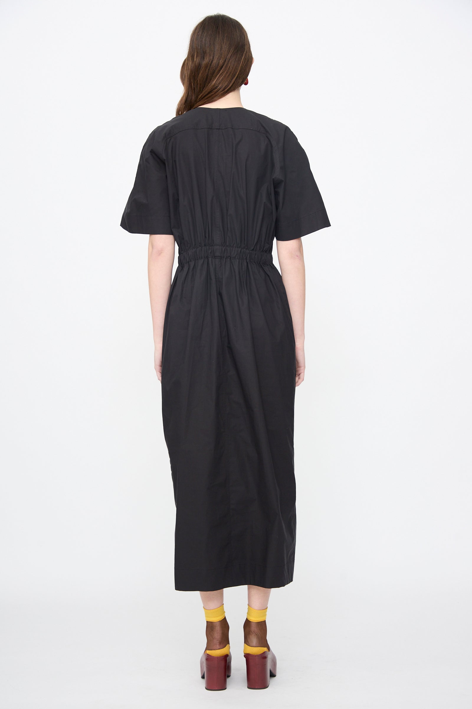 A person faces away wearing the Rachel Comey Ramses Dress in Black, a black midi dress with a gathered waist, made from organic cotton, paired with yellow socks and brown platform shoes against a plain white background.