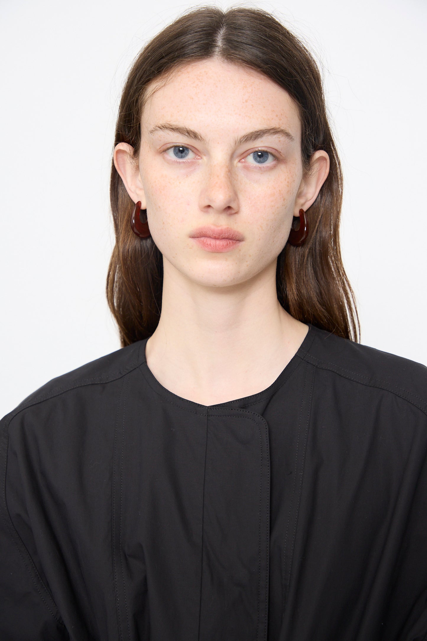 A young woman with long brown hair wears the Rachel Comey Ramses Dress in Black and red hoop earrings, posing against a plain white background.