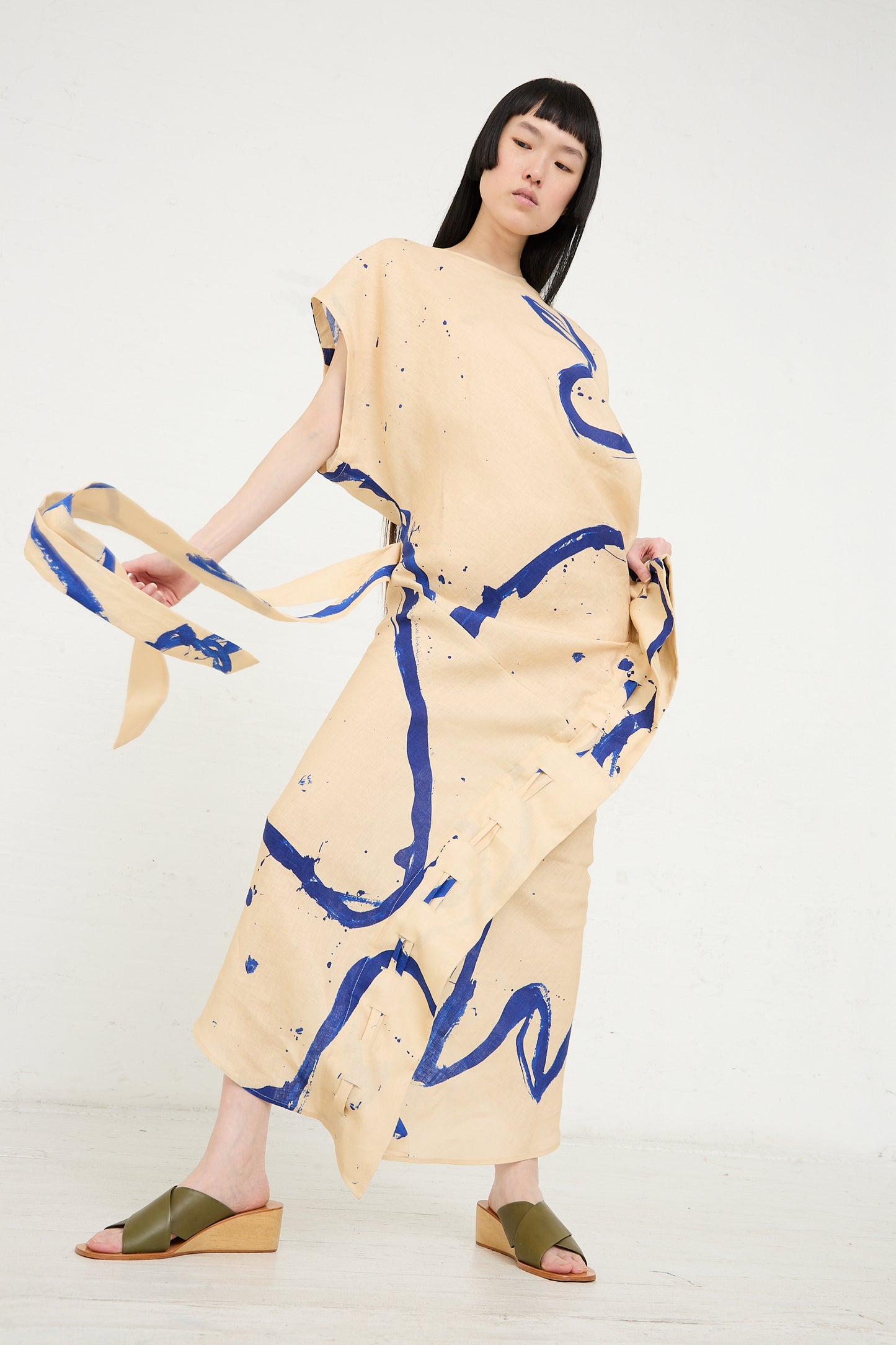 Woman posing in a Rachel Comey Regent Dress in Blush with blue abstract patterns, holding a matching belt, against a plain background.
