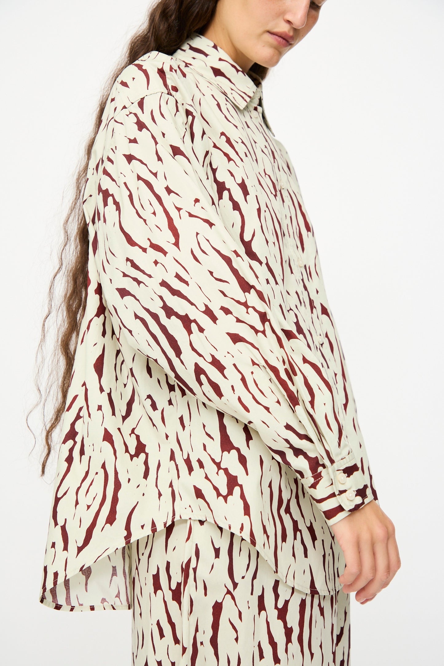 A person wearing the Rachel Comey Risa Top in Cream, a loose, long-sleeved cotton shirt with brown abstract Tiikeri print, paired with a matching skirt and standing against a plain background.