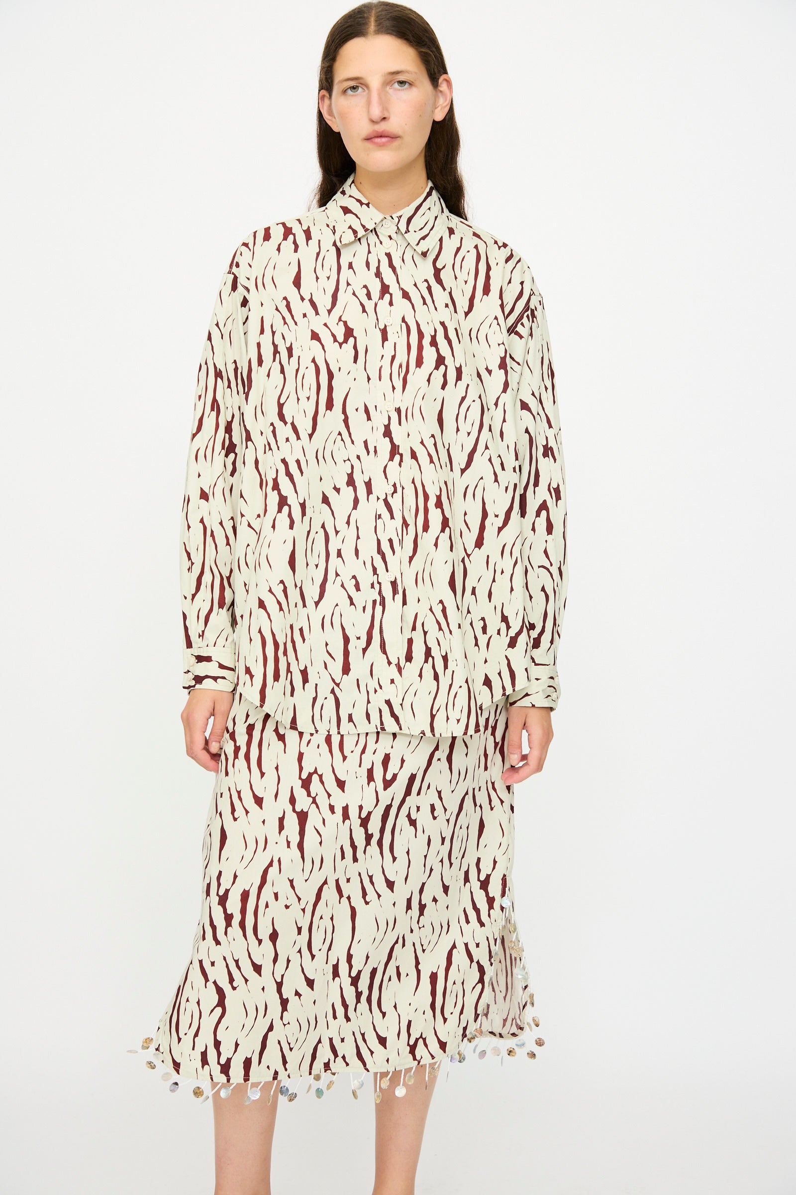A woman stands against a plain background wearing the Rachel Comey Risa Top in Cream, an oversized button-down shirt featuring an abstract beige and maroon pattern.