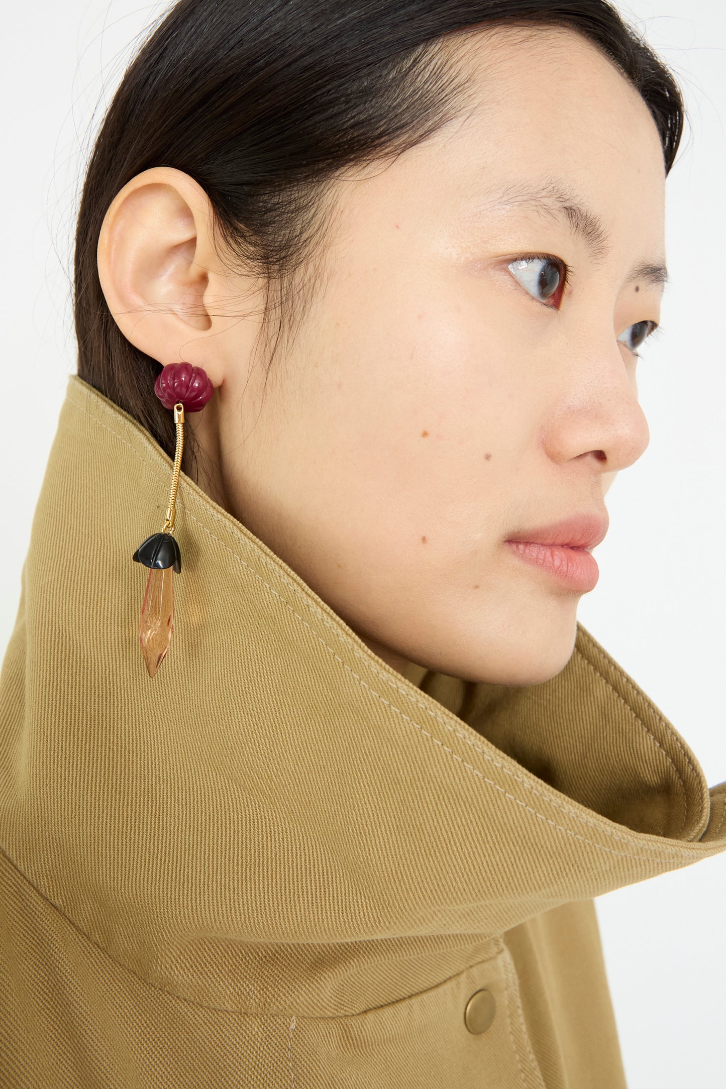 A person in a beige, high-collared coat showcases the Rosales Earring in Plum by Rachel Comey, featuring metal drop detailing in striking red, black, and brown hues.