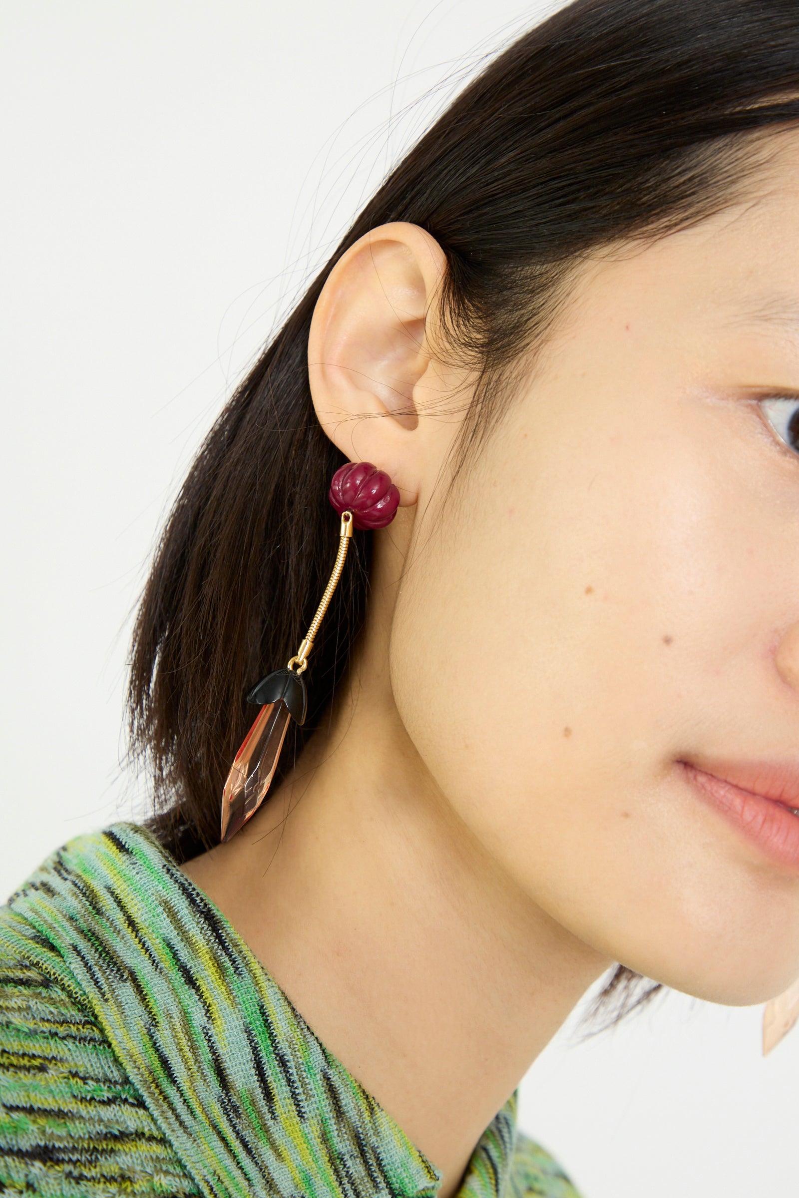 A person with short dark hair and a green patterned outfit wears the Rachel Comey Rosales Earring in Plum, showcasing burgundy, gold, black, and translucent elements with metal drop detailing.