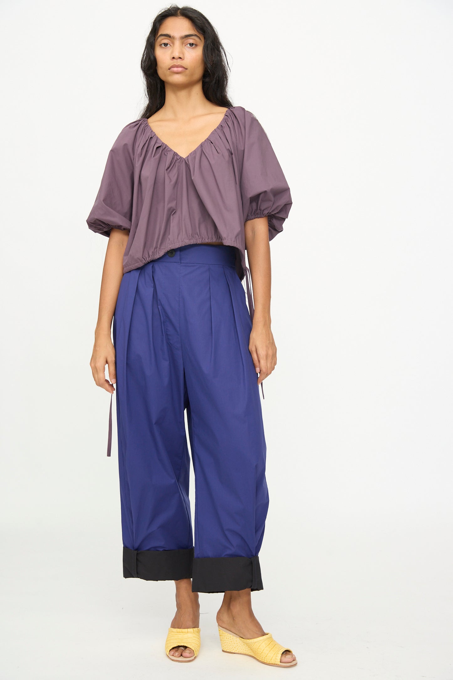 A person stands against a white background wearing the Shapley Top in Prune by Rachel Comey, blue wide-leg pants with black cuffs, and yellow sandals.