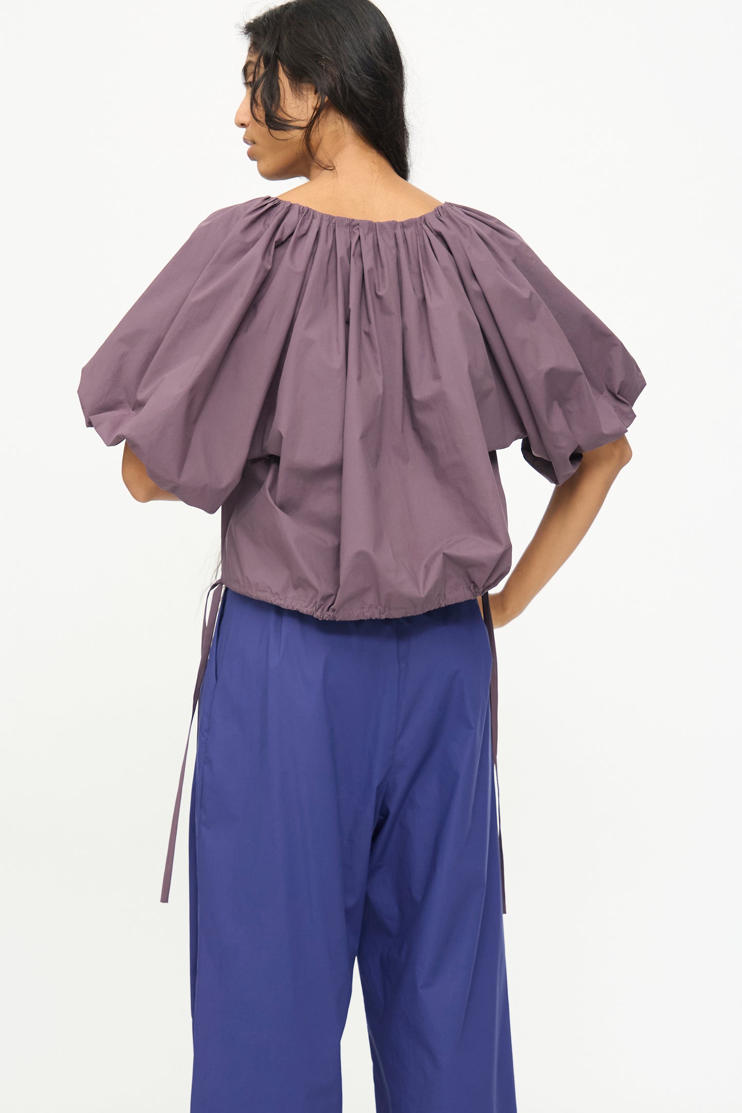 A person stands turned away, wearing the Rachel Comey Shapley Top in Prune with wide blue pants, set against a plain white background.