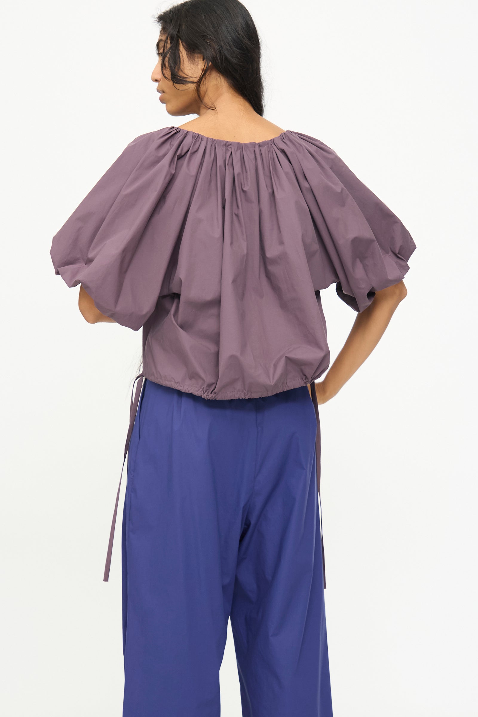 A person stands turned away, wearing the Rachel Comey Shapley Top in Prune with wide blue pants, set against a plain white background.