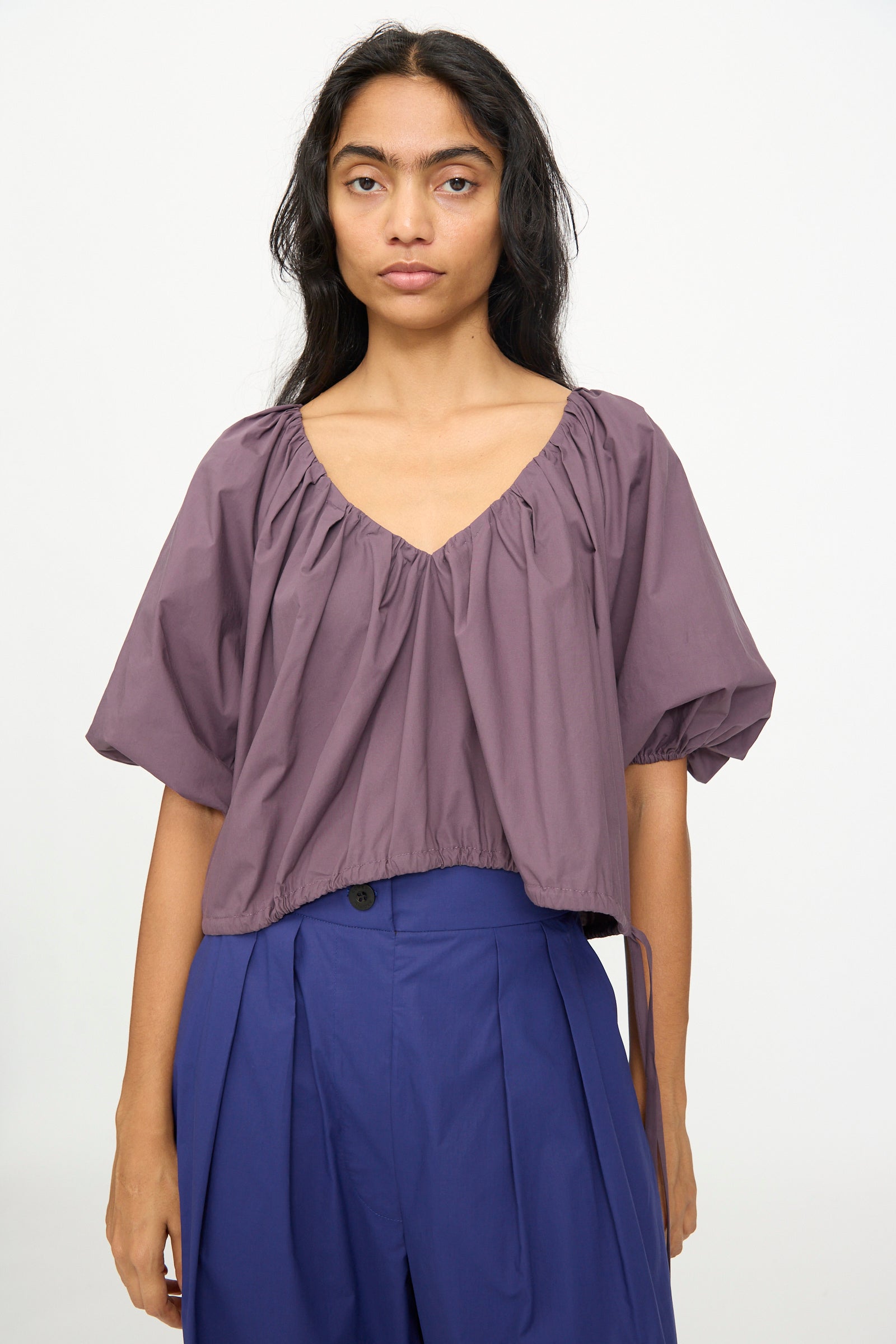 A person with long dark hair wears the Rachel Comey Shapley Top in Prune, an organic cotton blouse with a gathered neckline and short sleeves, paired with high-waisted blue pleated pants, standing against a plain light background.