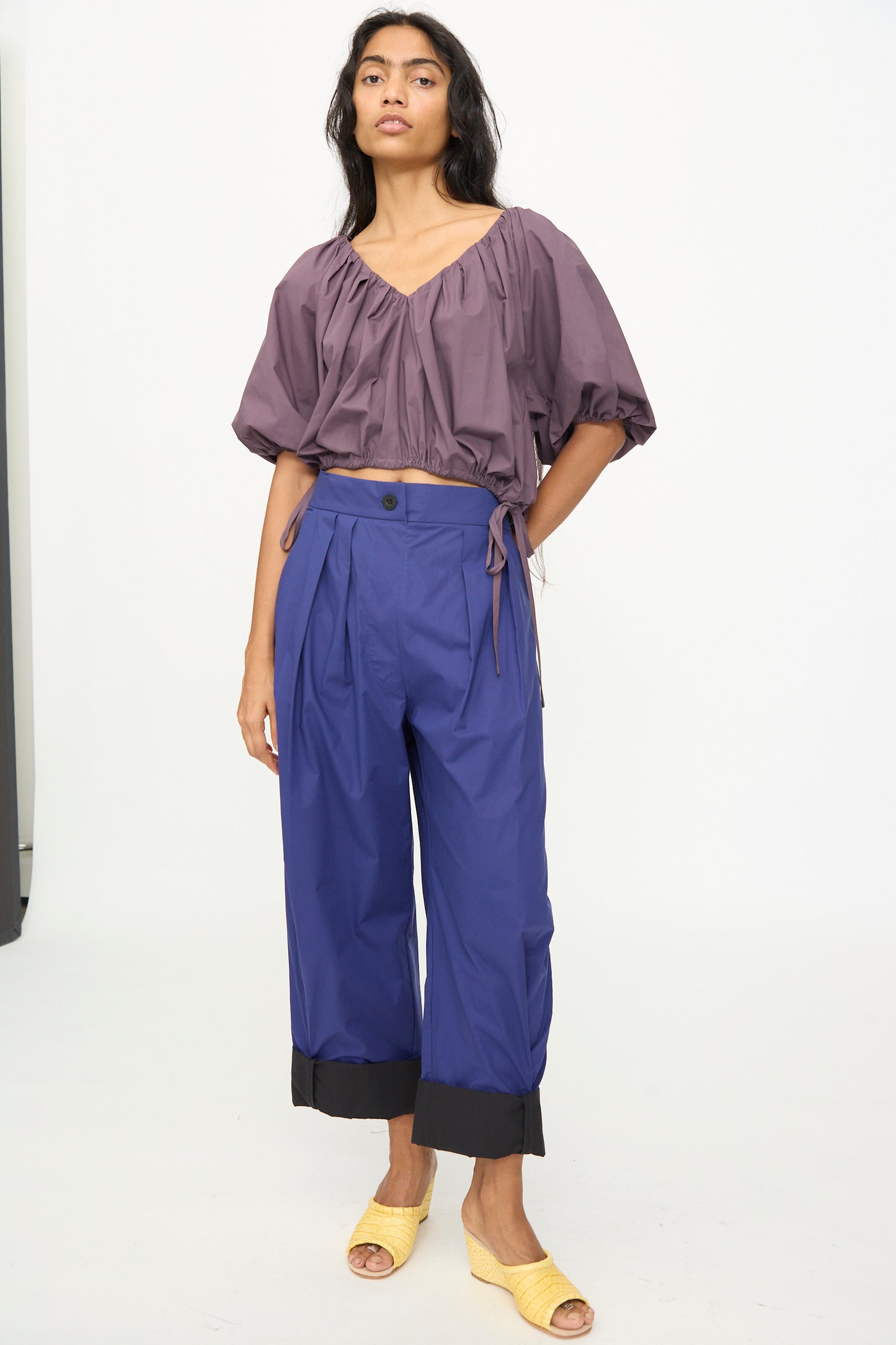 A person stands against a white background wearing the Rachel Comey Shapley Top in Prune, loose purple balloon sleeve blouse, wide blue pants with black cuffs, and yellow open-toed shoes.