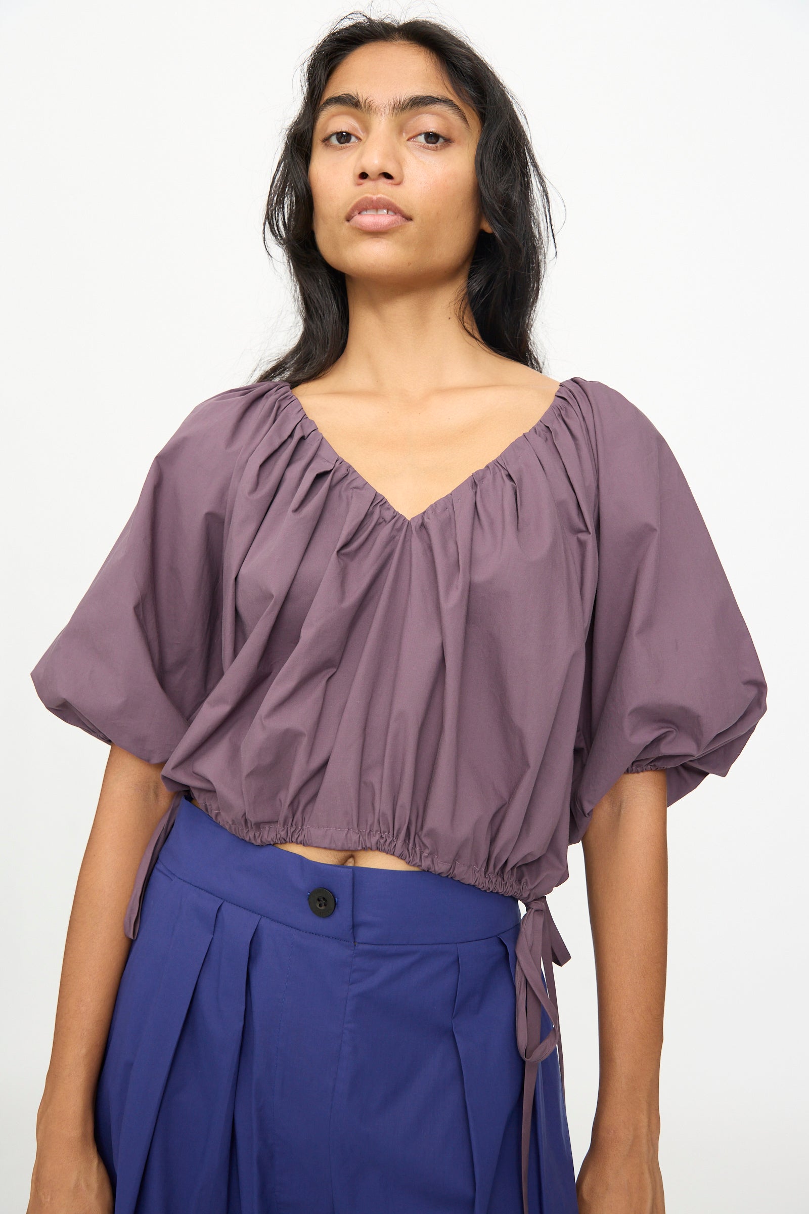 A person wearing the Shapley Top in Prune by Rachel Comey—a loose-fitting, short-sleeved organic cotton top with a gathered neckline—paired with blue high-waisted pleated pants, standing against a plain light background.
