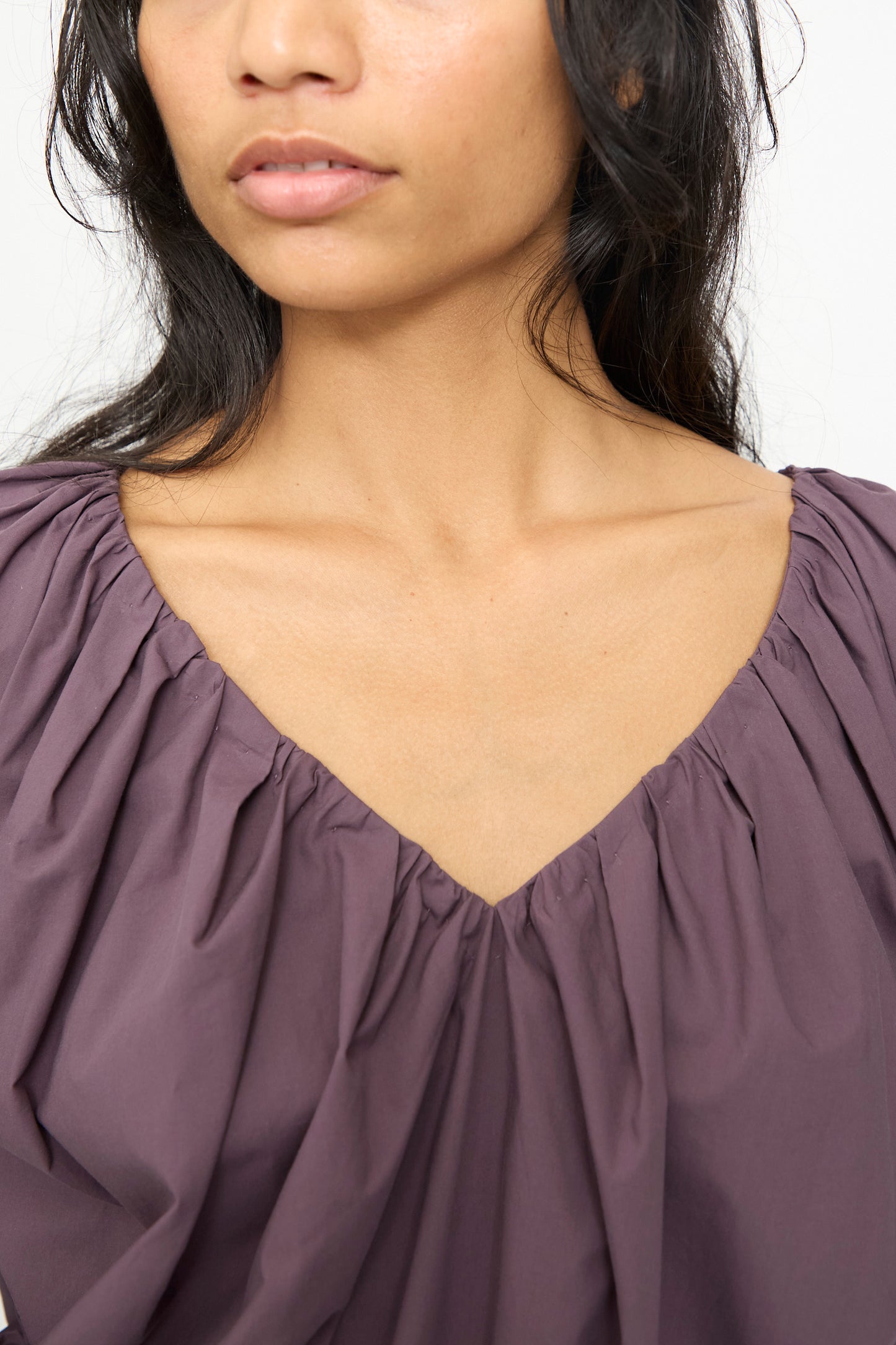 A woman wearing the Rachel Comey Shapley Top in Prune, an organic cotton piece with a gathered V-neck. The image centers on her collarbone and shoulders, showing just her lower face.