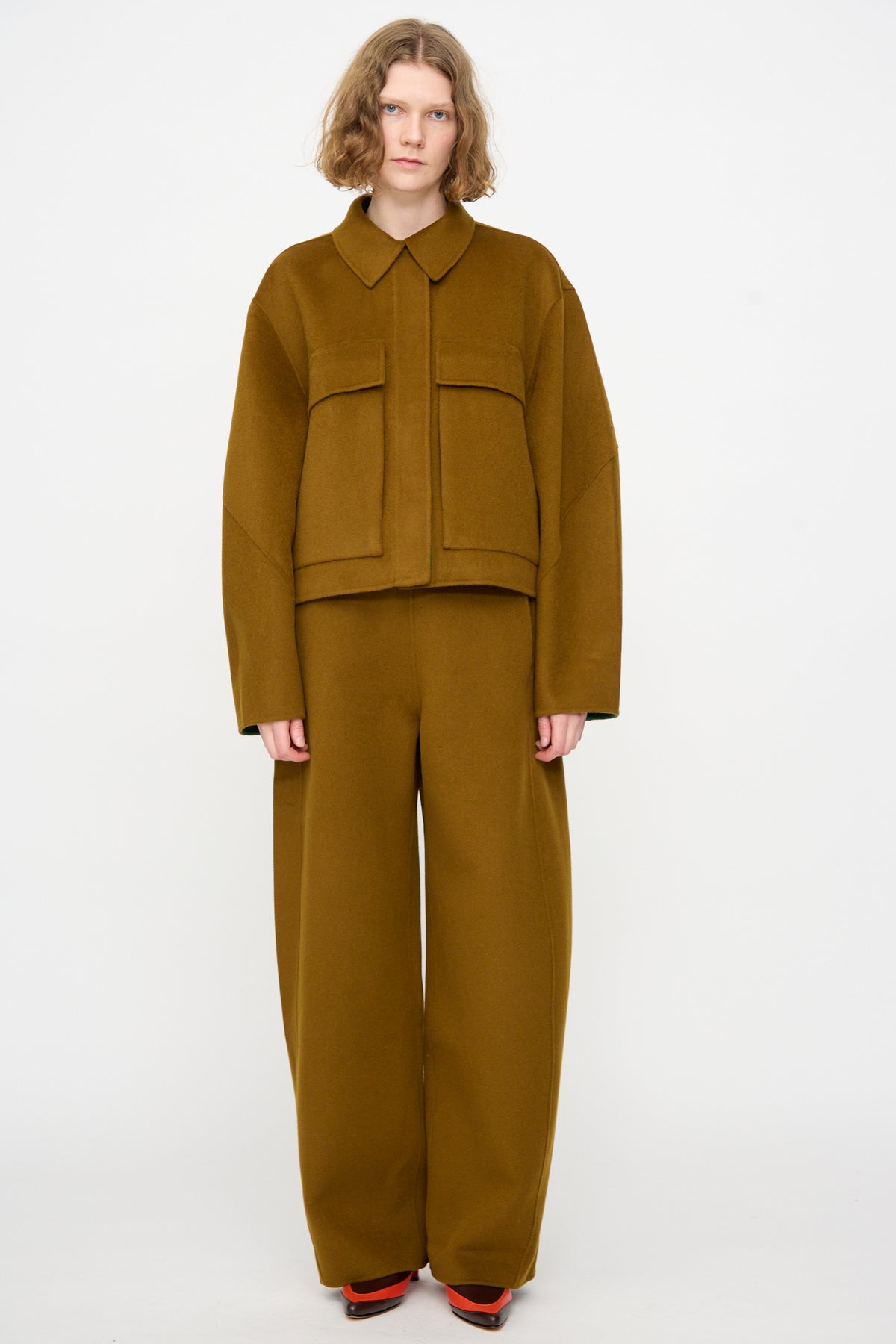 A person wearing the Rachel Comey Site Jacket in Bronze—a loose-fitting, double-faced wool button-up with large pockets—paired with matching wide-leg pants, stands against a plain white background.
